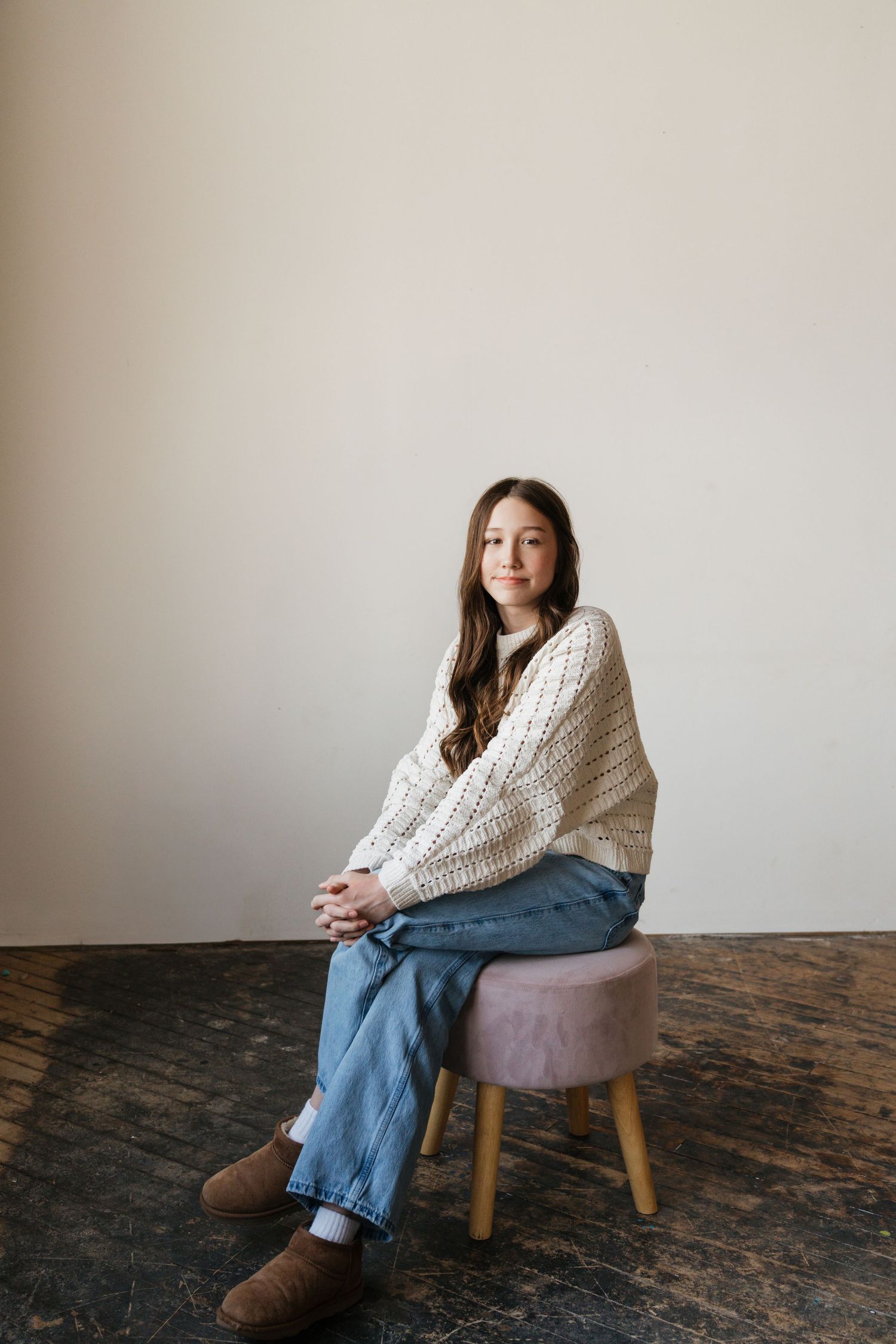 A teenage girl in a cream sweater and jeans sits on a pink stool against a white wall.
