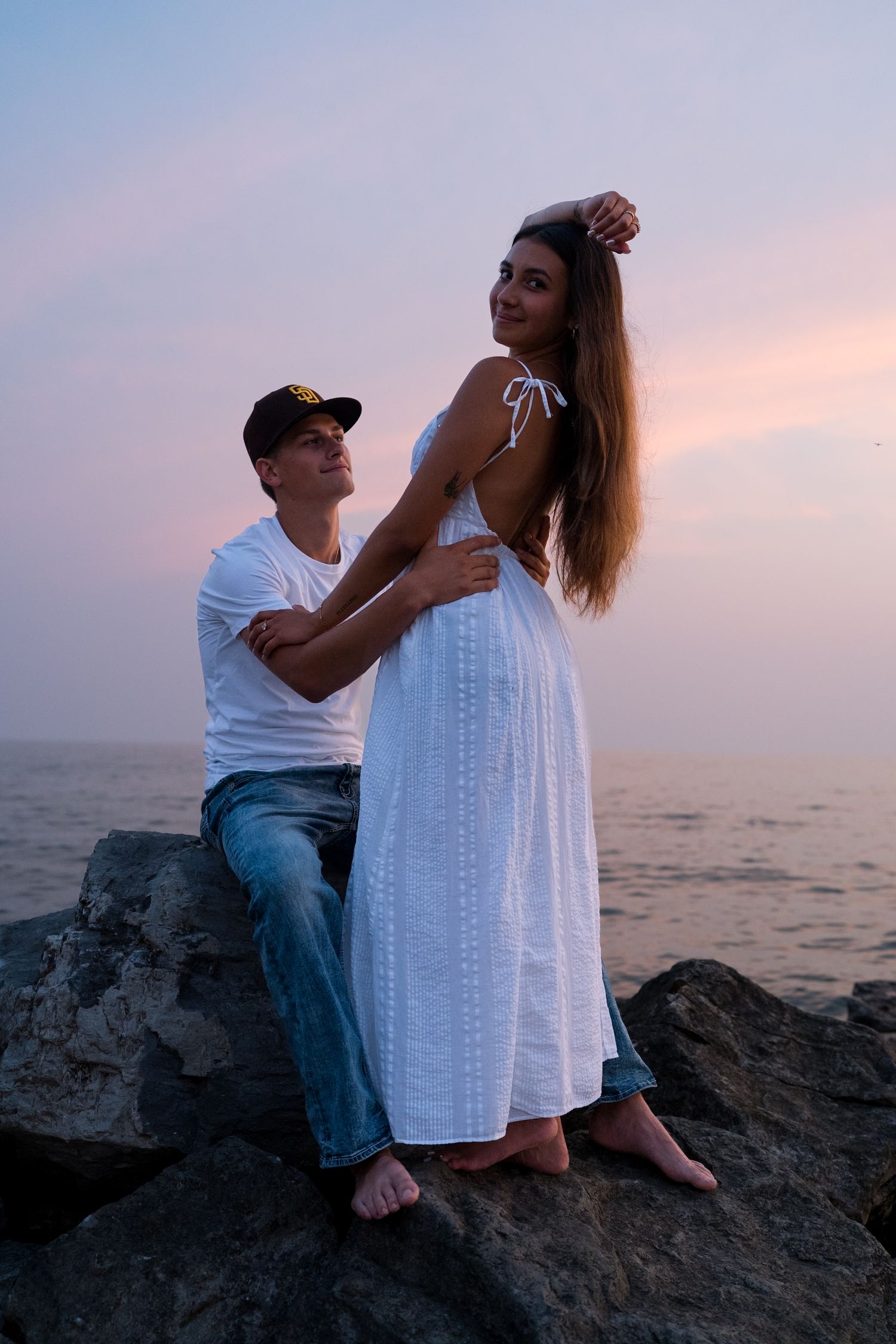 Young couple posing on rocks by ocean at dusk, man embracing woman in backless white maxi dress at sunset.