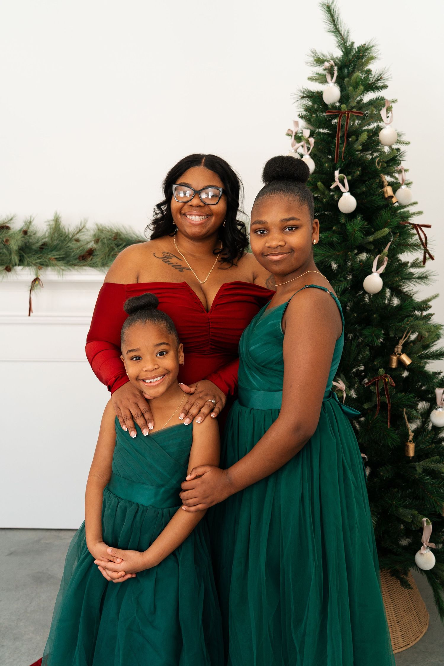 Mother in red off-shoulder top poses with two daughters in green tulle dresses by Christmas tree.