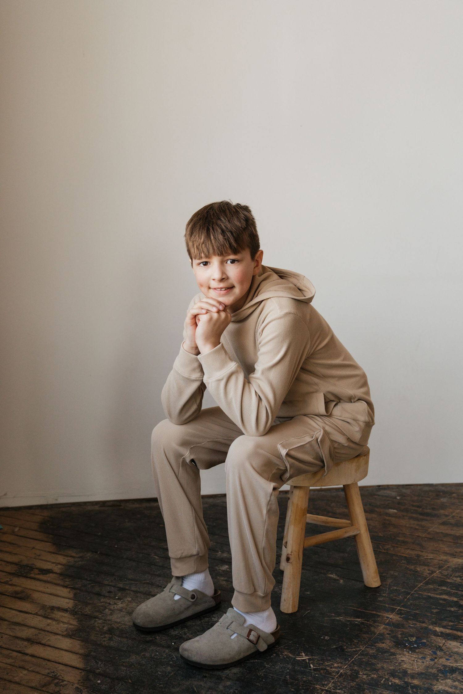 A smiling boy in a beige hoodie tracksuit sits on a wooden stool against a white wall.