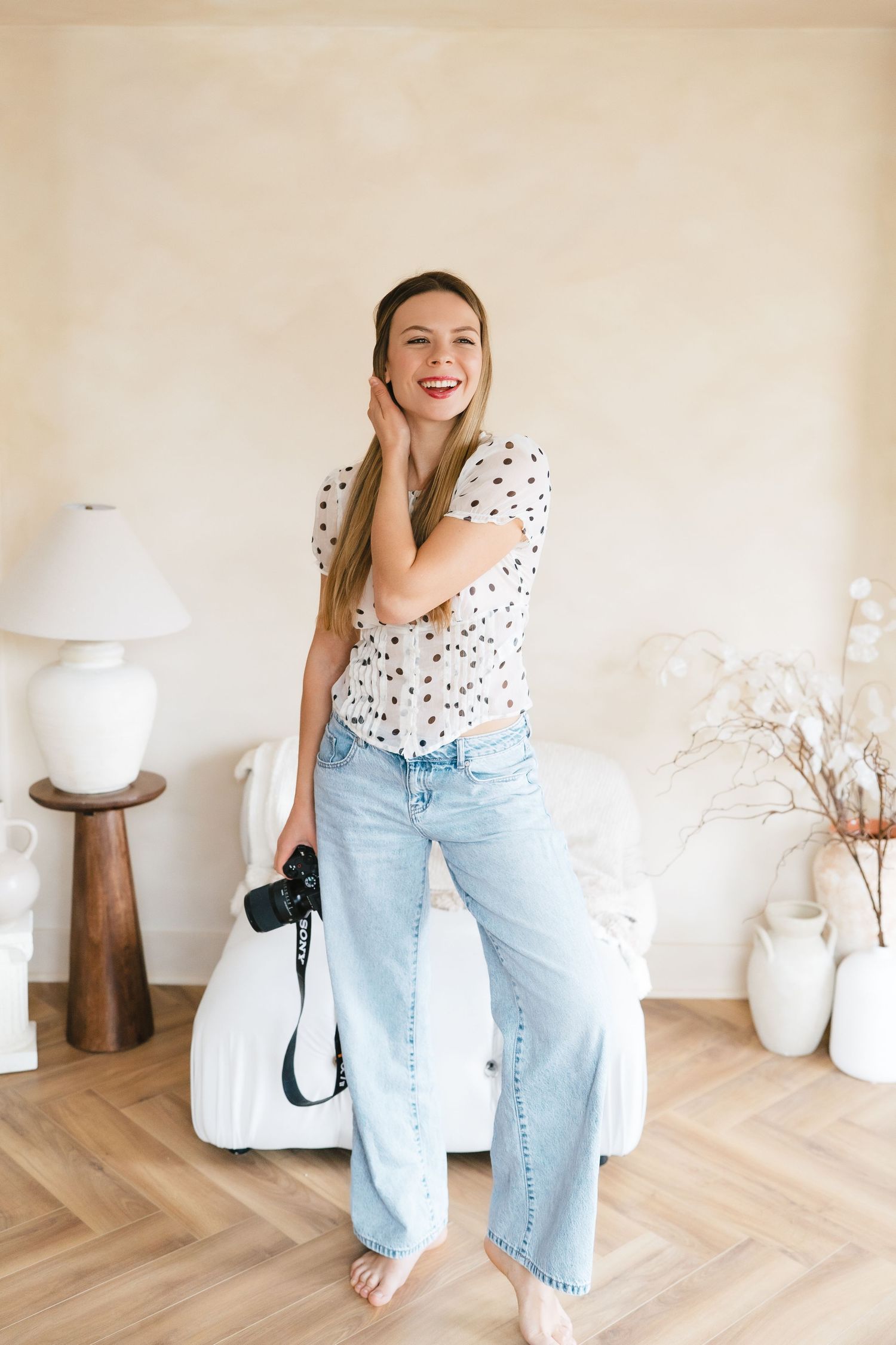 Laughing woman in polka dot top and light jeans holding camera while standing in bright modern interior.
