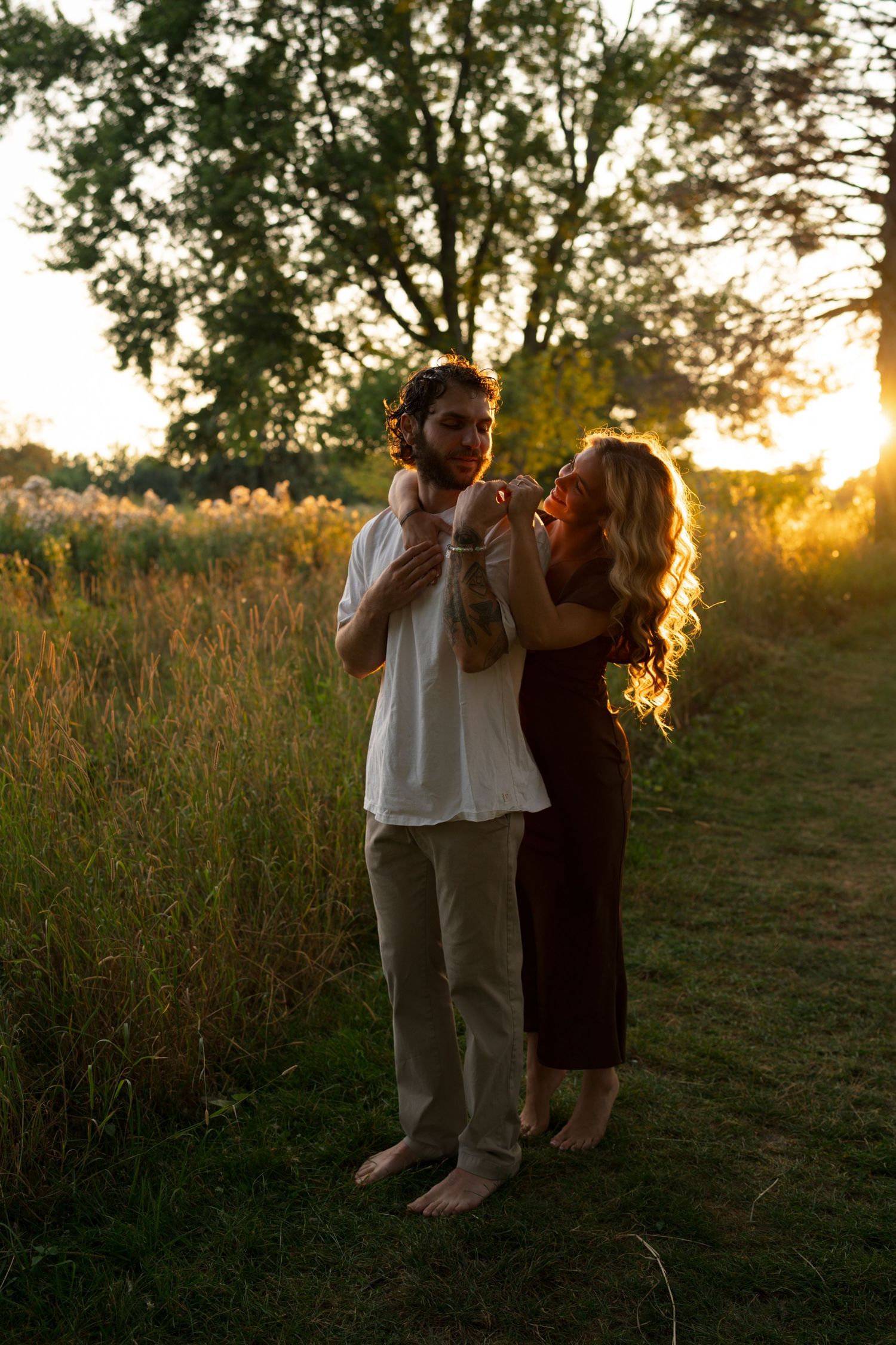 Couple standing barefoot in golden sunset field, woman in black dress touching man in white shirt near trees.