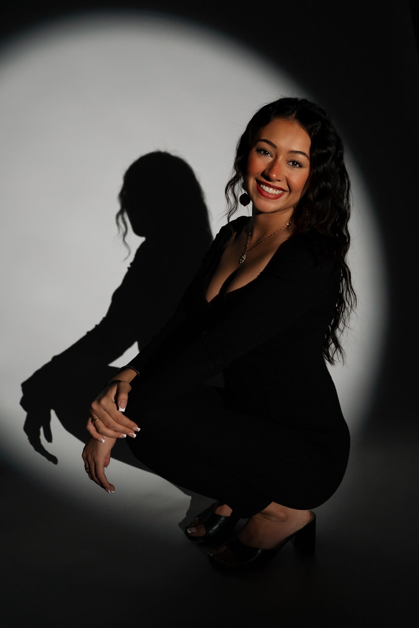 Confident woman in black outfit crouching and smiling in a dramatic circular spotlight portrait.