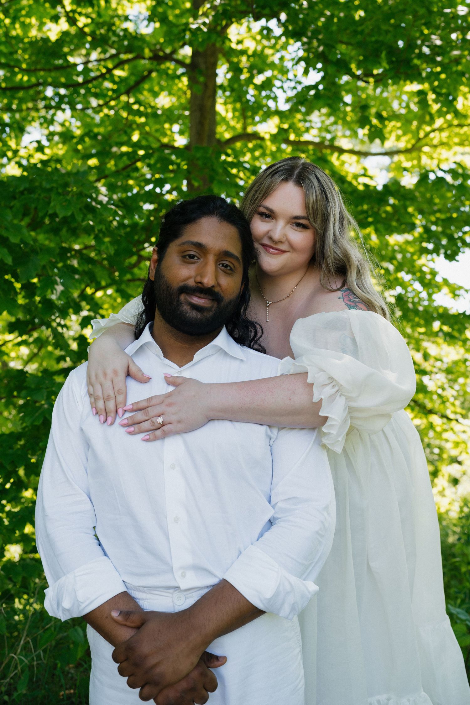 Interracial couple posing together outdoors under green trees, woman in white dress embracing man.