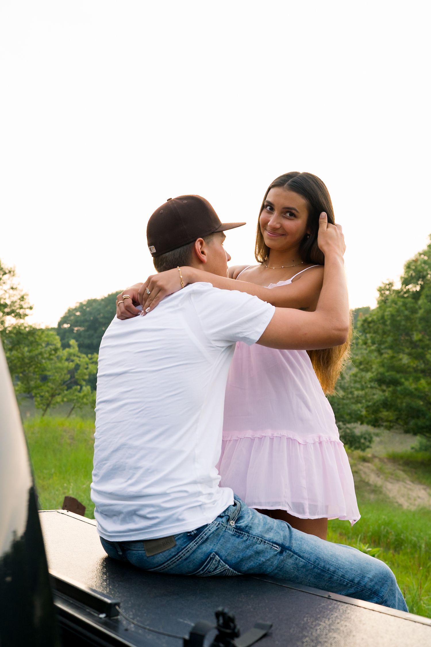 Young couple embracing outdoors near truck at sunset, woman in pink dress hugging man in white tee and cap.