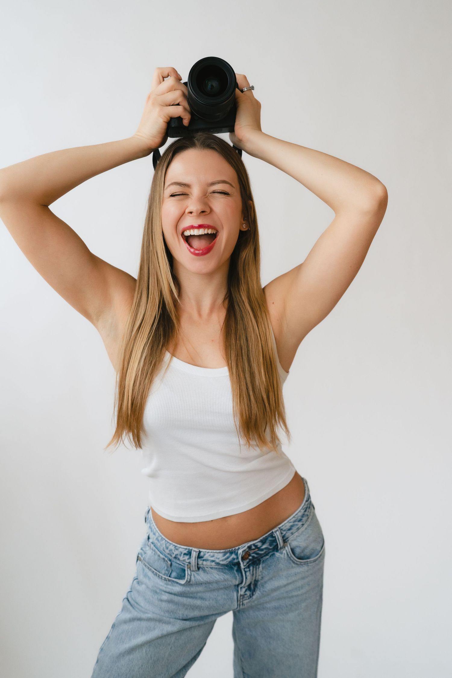 Excited young blonde woman in white tank top holding camera overhead against white studio background.