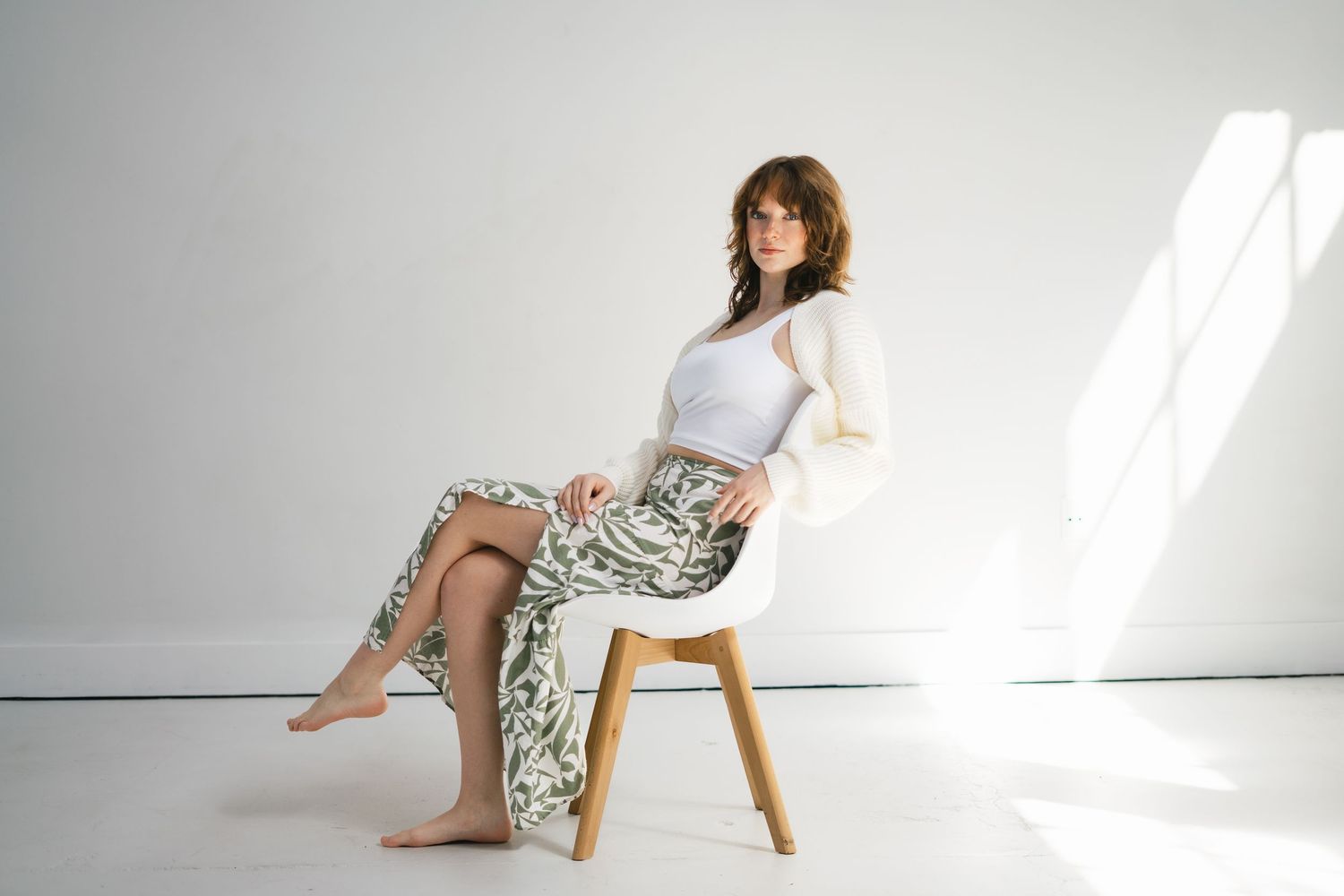 Woman in white top and sage green floral maxi skirt sitting barefoot on modern white chair in studio.