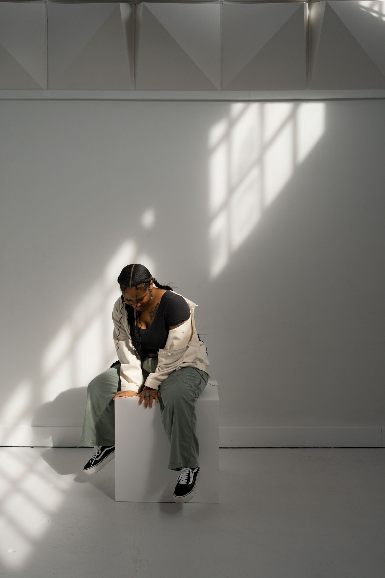 A person in sage green pants and white jacket sits on a white cube in a sunlit minimalist white studio space.