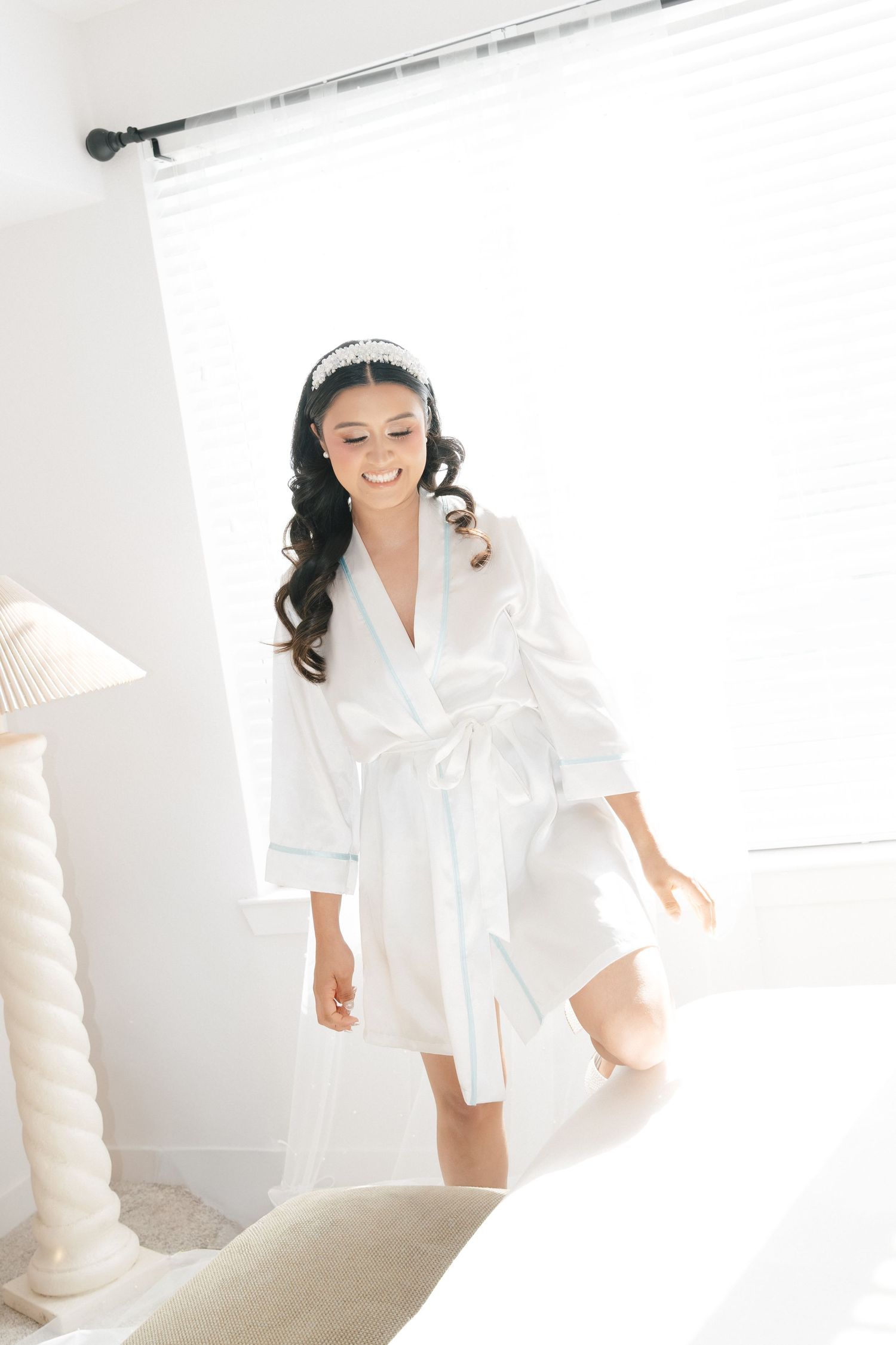 Smiling bride in white satin robe and pearl headband getting ready on her wedding day.