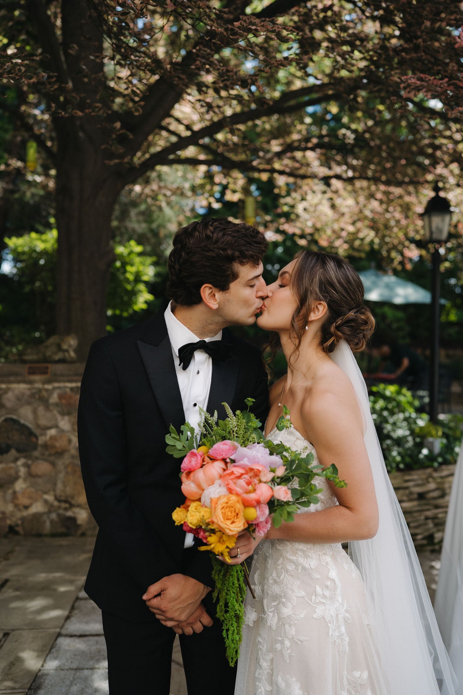Bride and groom share a romantic kiss outdoors, bride holding a vibrant pink and orange floral bouquet.