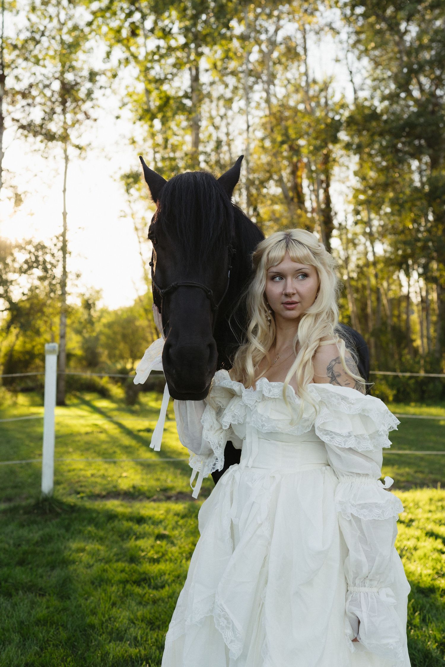 Blonde woman in white wedding gown poses with a dark horse on green grass surrounded by trees.