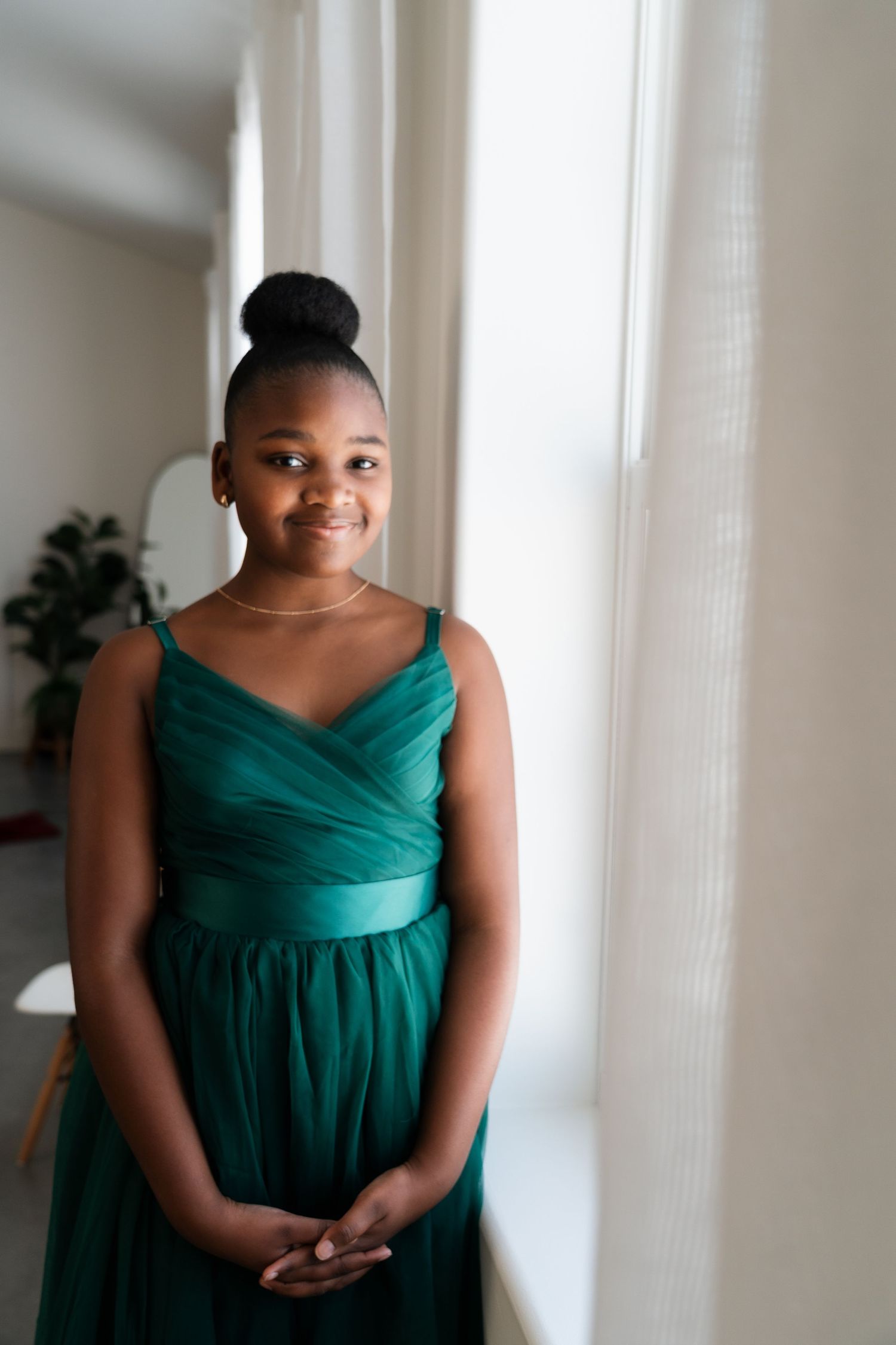 Young girl in elegant green tulle gown stands by a bright window with hands clasped together.