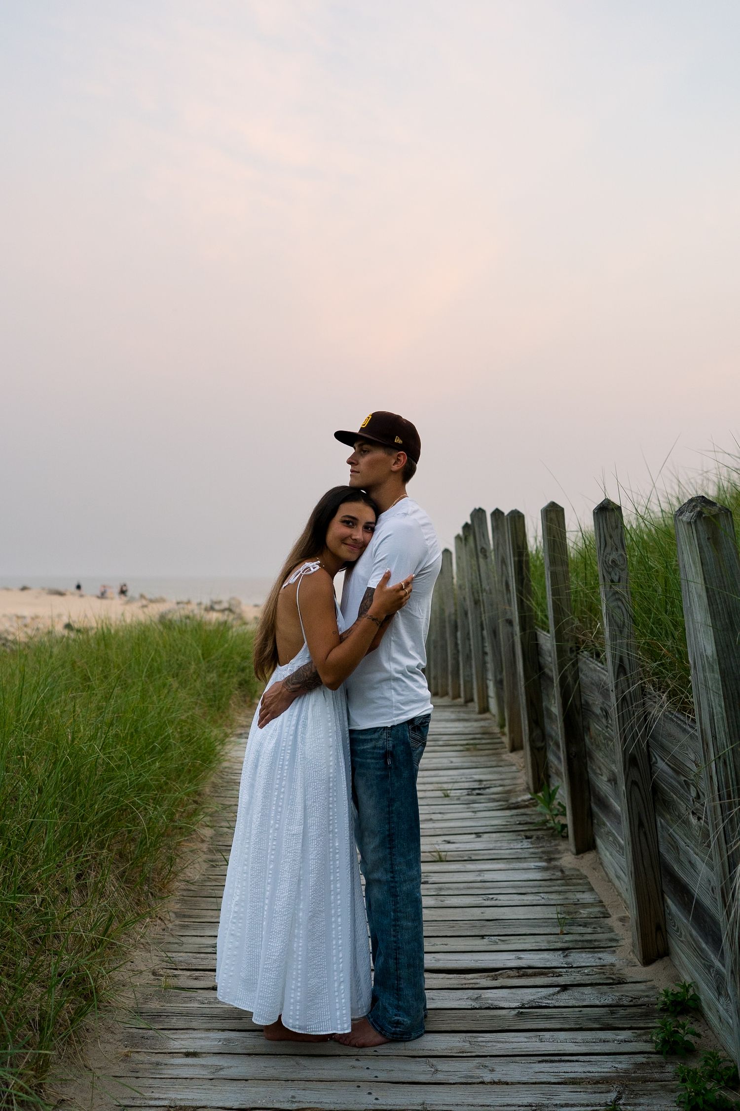 Romantic young couple embracing on wooden beach boardwalk path at sunset, woman in white dress, man in jeans.