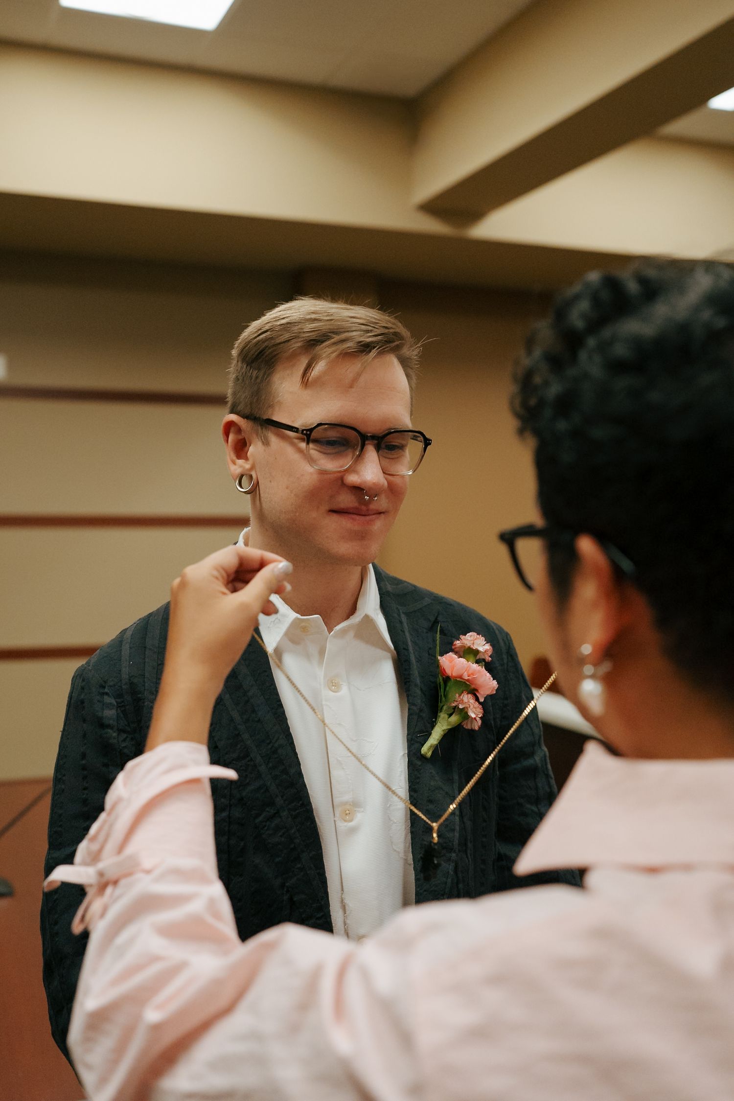 Spouse placing necklace on partner in dark blazer with pink boutonniere at courthouse wedding.