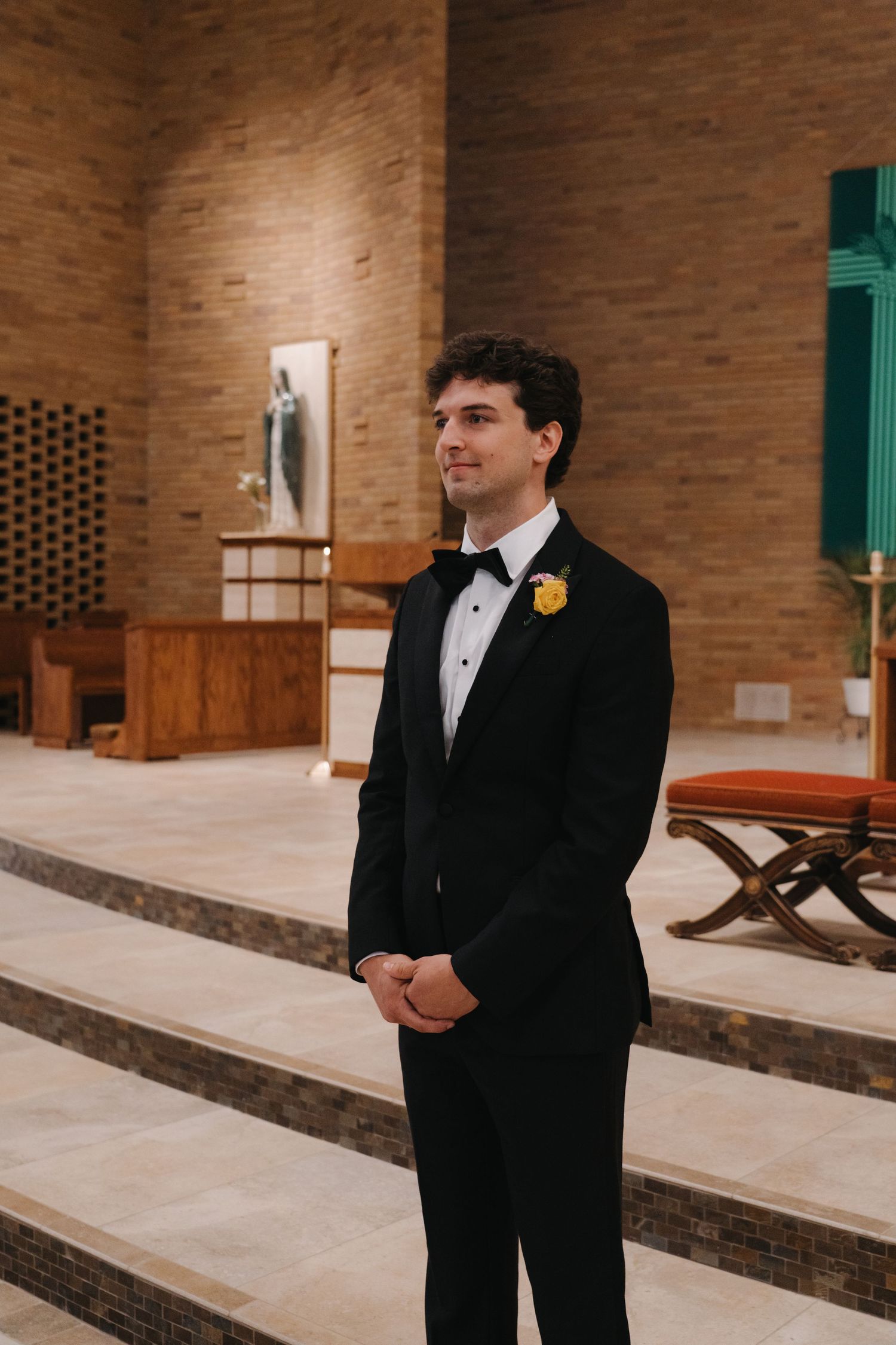 Handsome groom in black tuxedo with yellow boutonniere stands at church altar awaiting his bride.