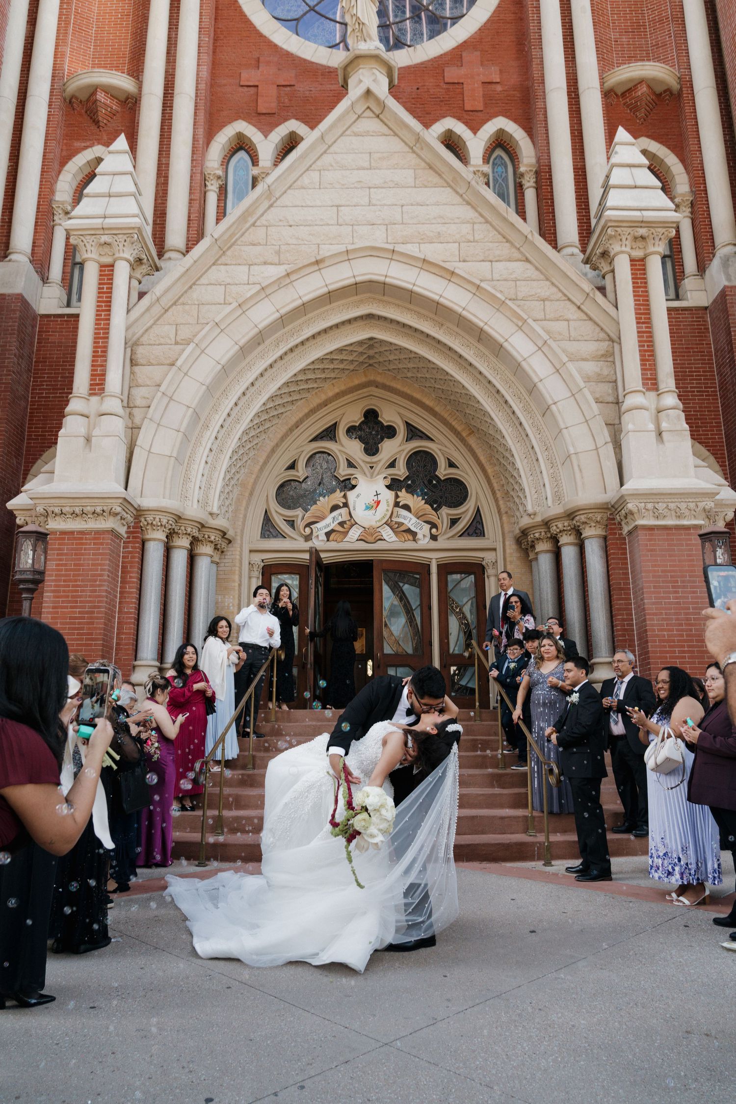 Groom dips bride for a romantic kiss on the steps of a grand red brick Gothic church.