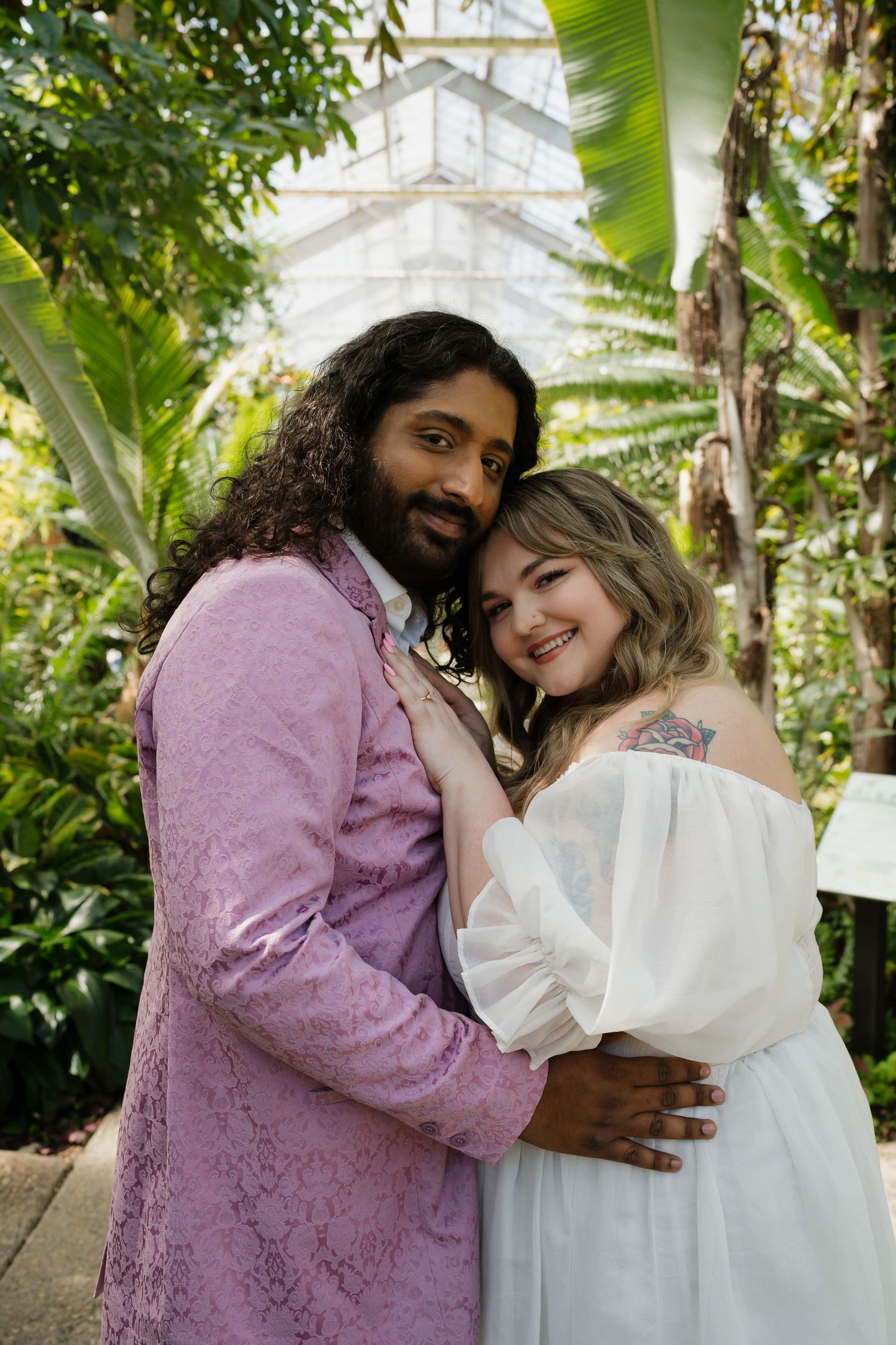 Smiling interracial couple embracing in tropical greenhouse, groom in pink lace suit.