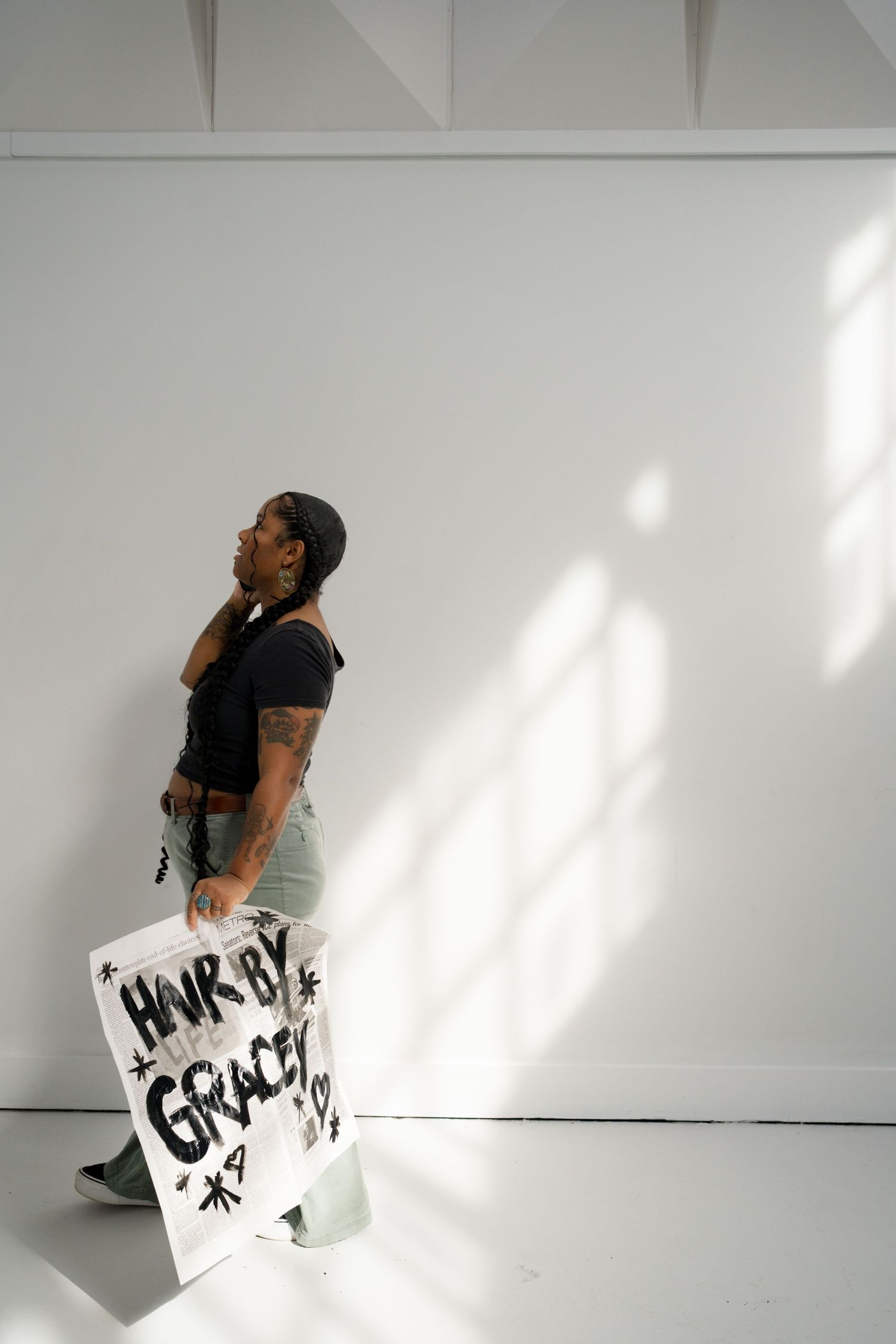A Black woman with braids walks through a bright white studio carrying a sign reading 'Honk By Grace' in bold black letters.