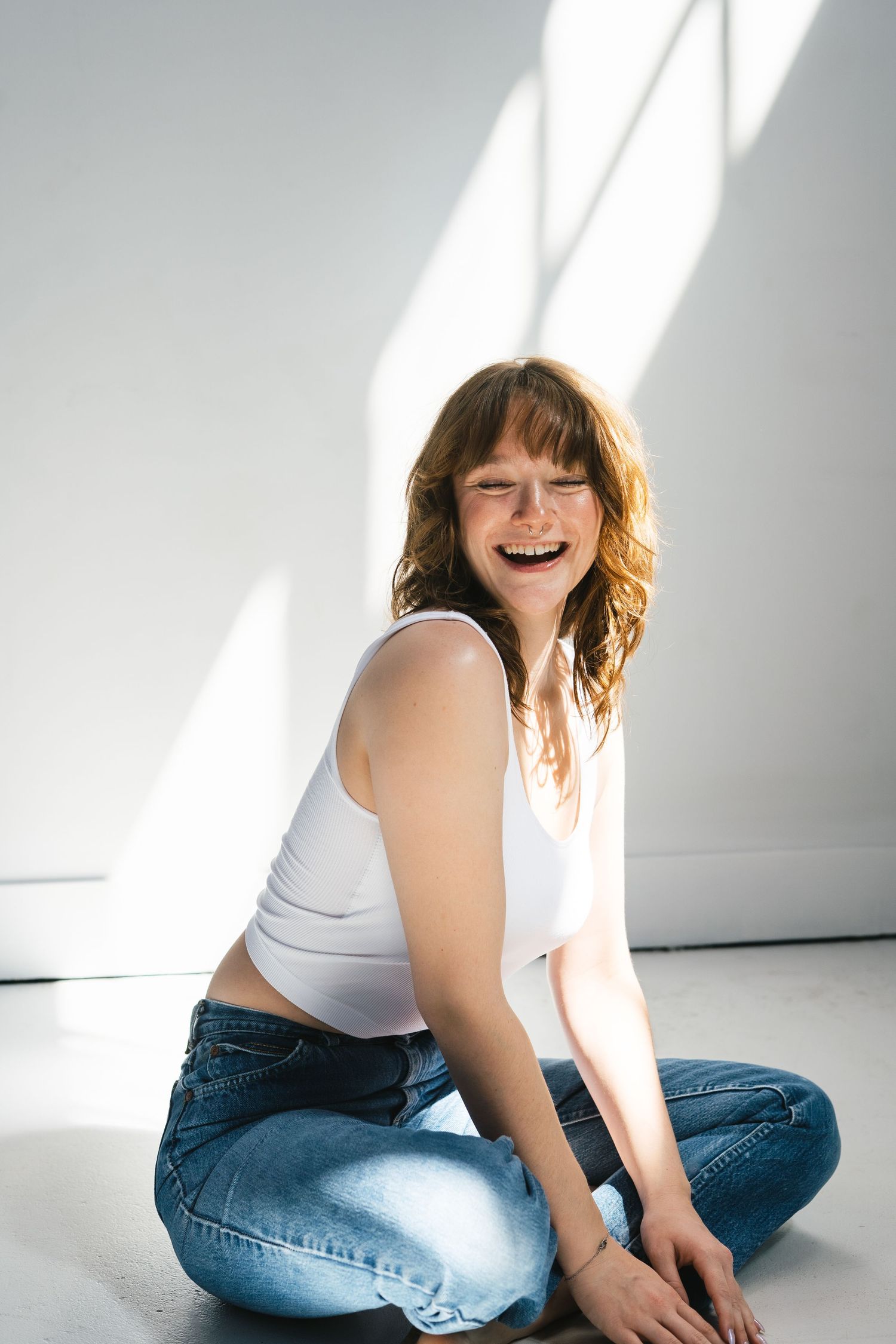 Laughing woman in white tank top and blue jeans sitting cross-legged on floor in bright sunlit studio.