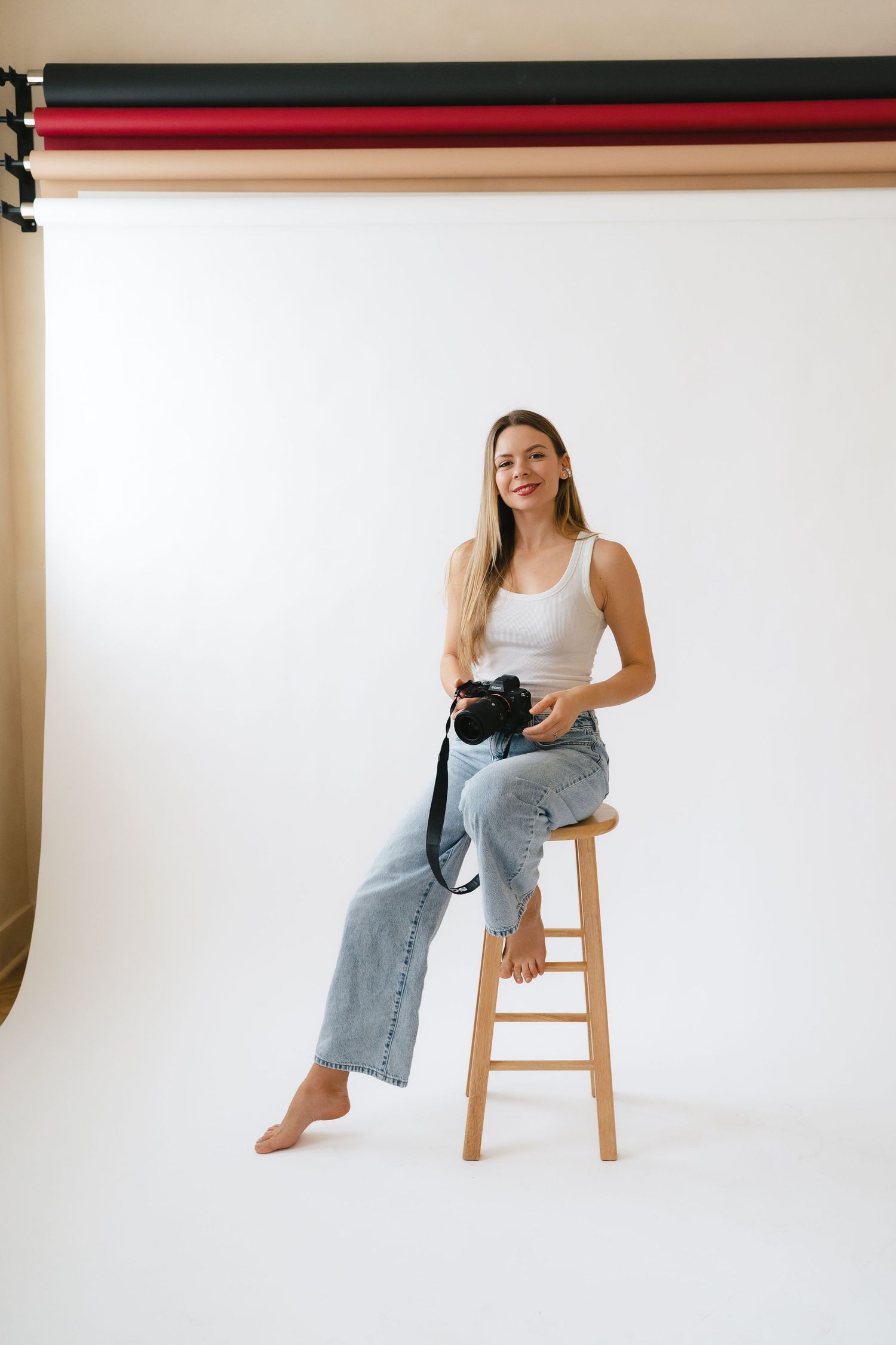 Smiling female photographer in white tank and jeans sitting on stool holding camera in professional photo studio.