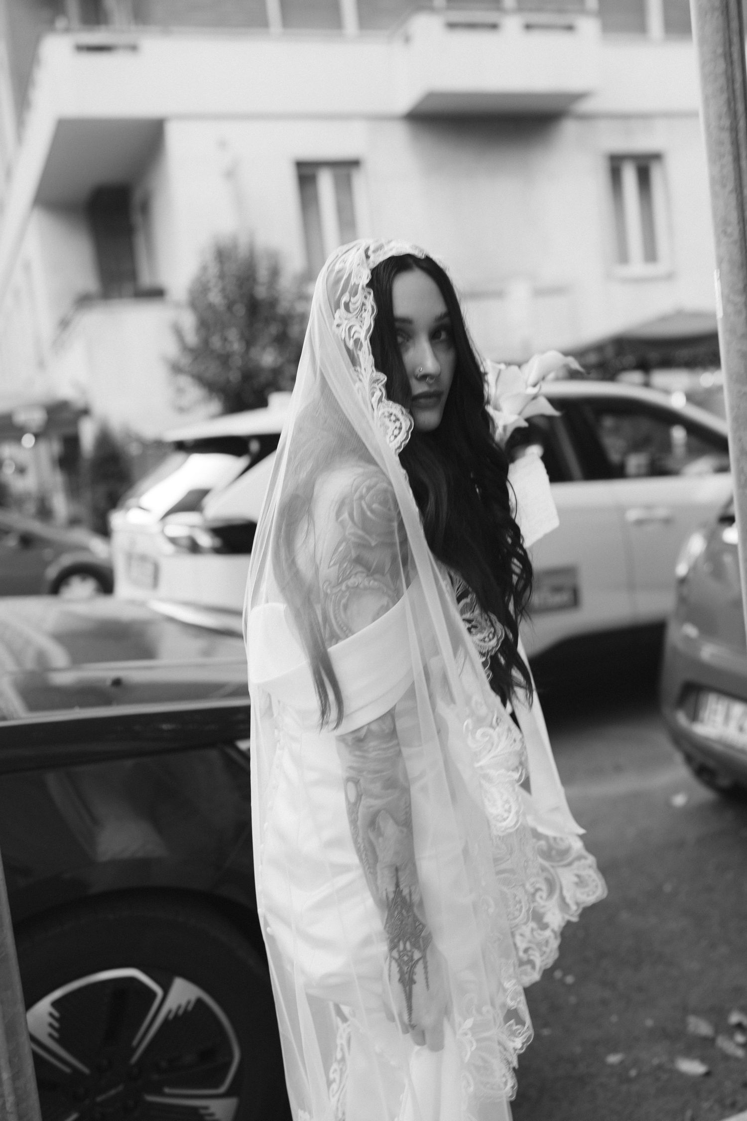 Black and white photo of tattooed bride in lace veil holding flower, standing near cars on urban street.