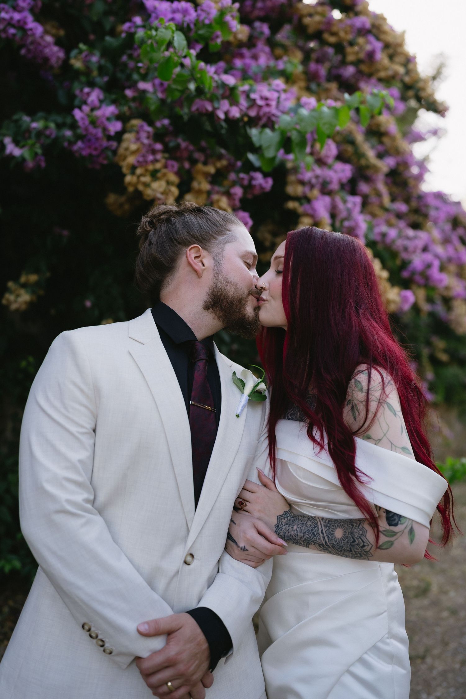 A couple shares a romantic kiss at their wedding, with the groom in a white suit and bride with red hair before purple blooms.