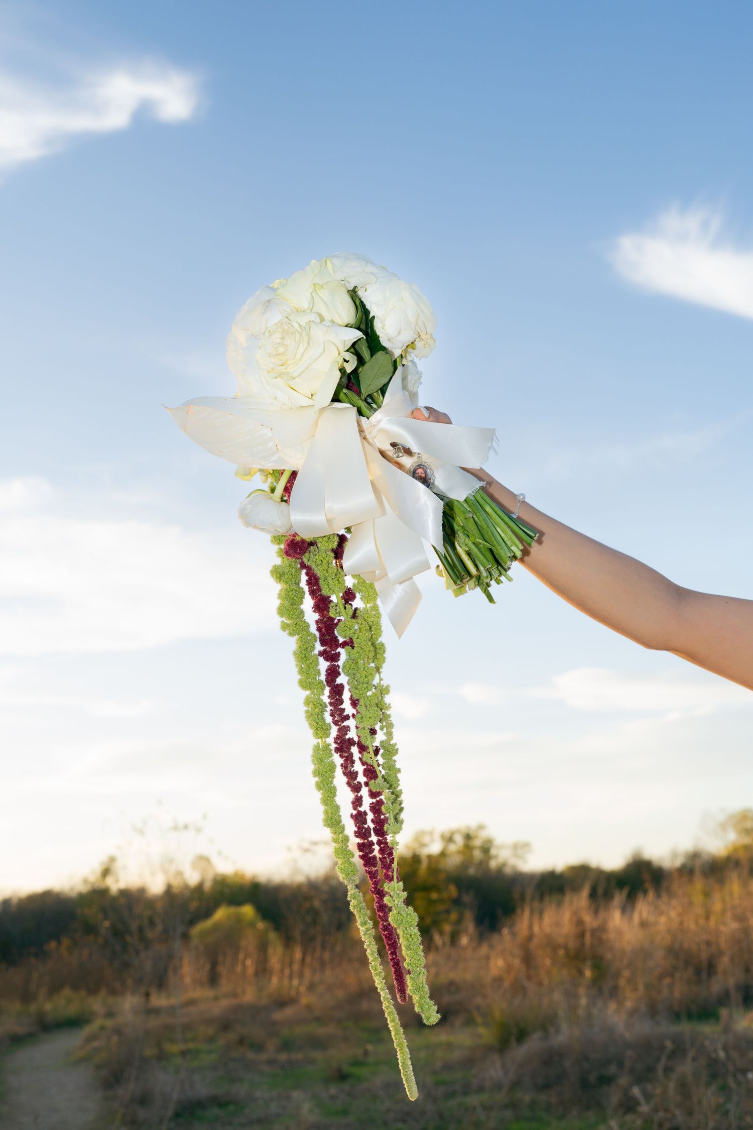 Hand holds a white peony bouquet with trailing burgundy and green amaranthus against a blue sky.