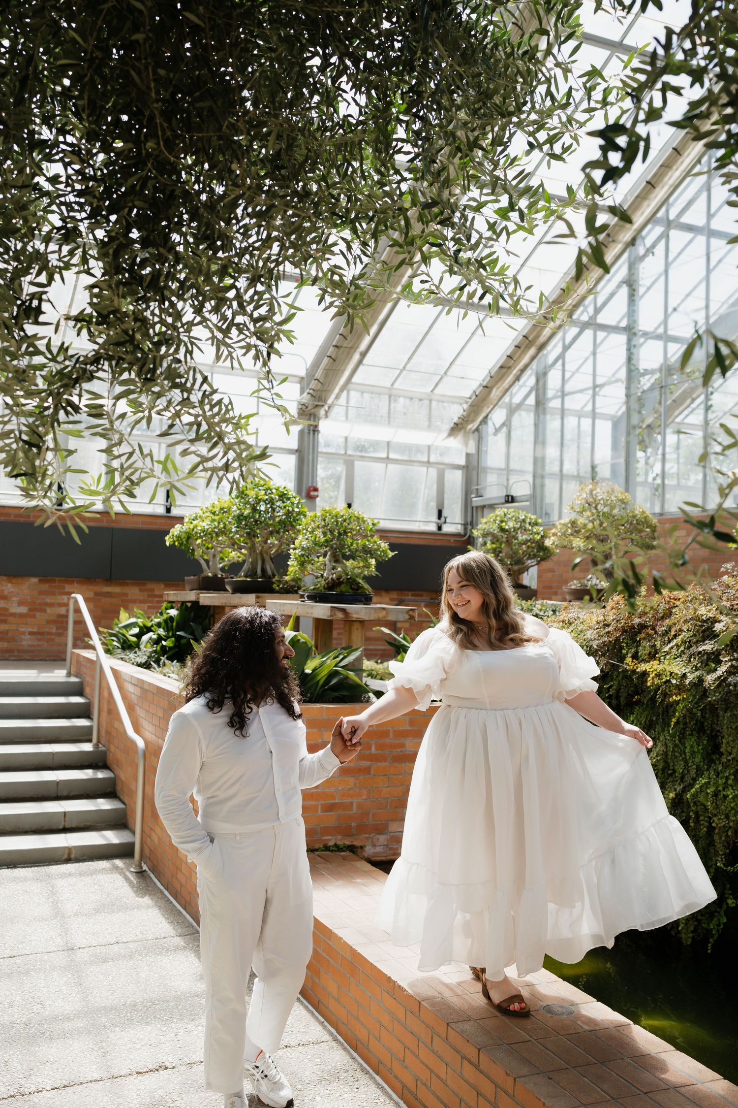 Bride in white dress holds partner's hand while dancing in sunlit botanical garden greenhouse.