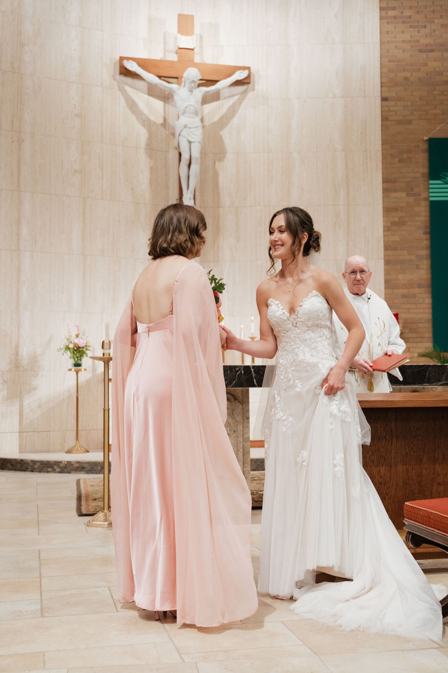 Bride in lace gown holds hands with bridesmaid in pink dress at the church altar during ceremony.