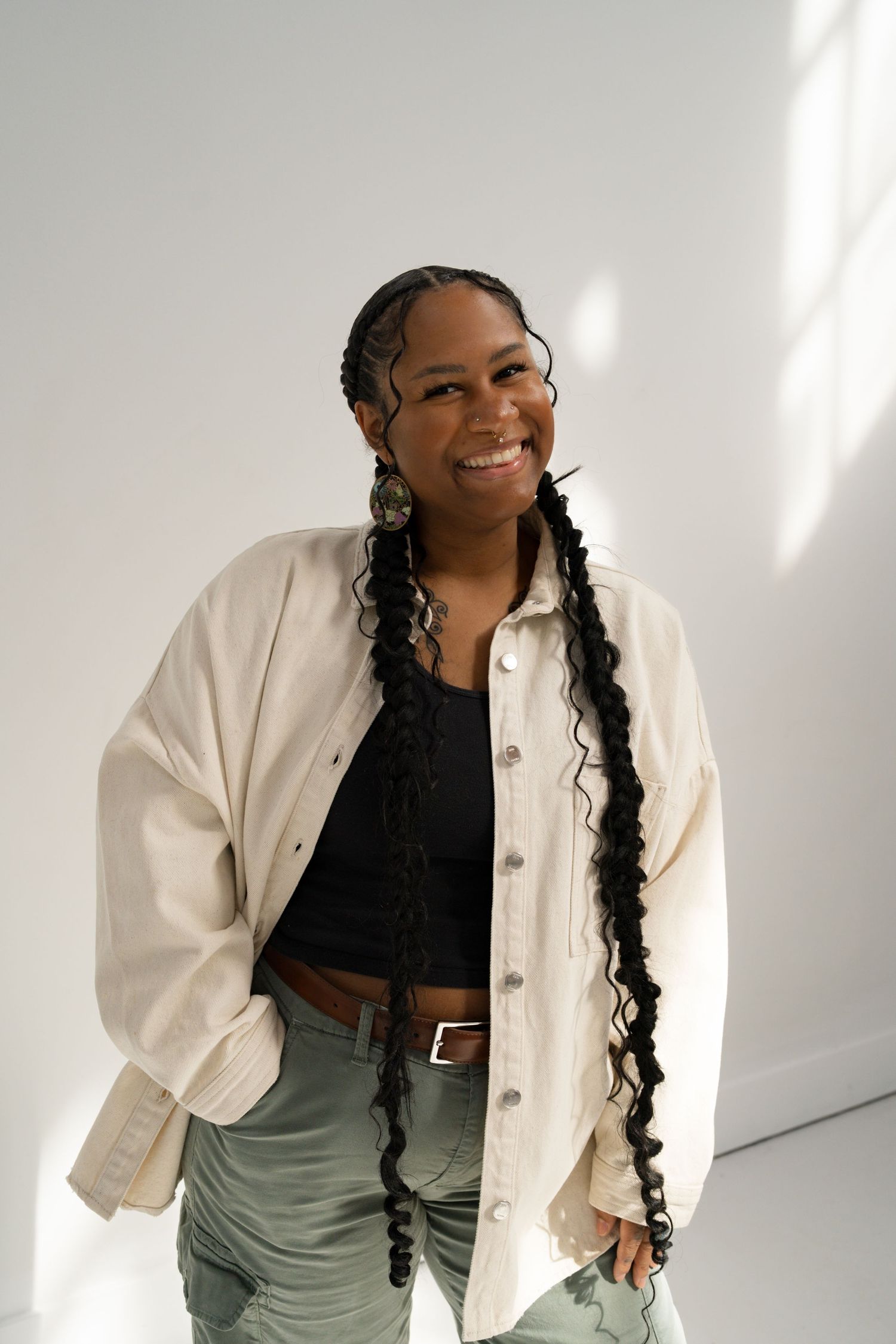 A smiling Black woman with long braids poses in a white jacket, black crop top, and sage green pants against a white wall.