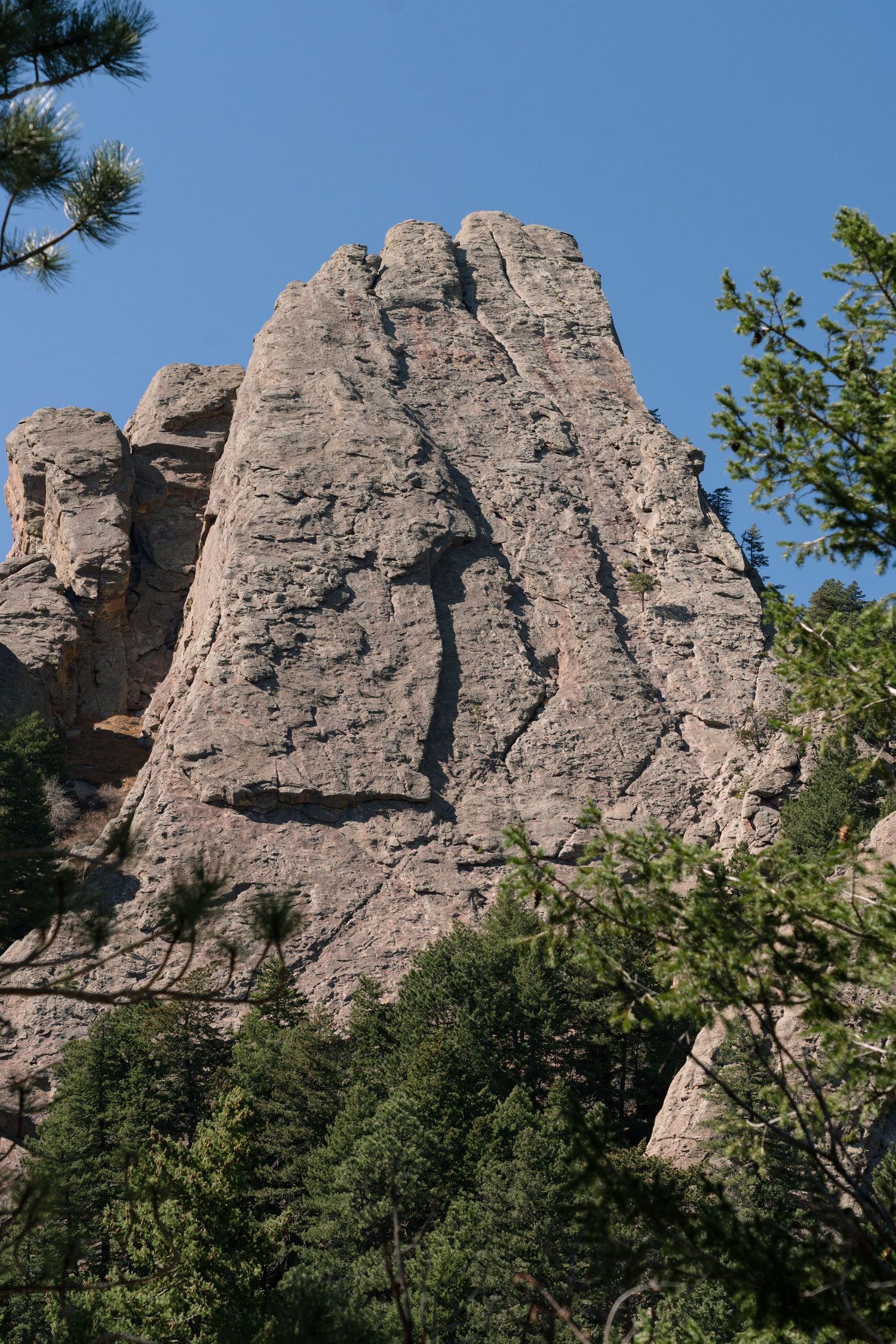 A dramatic rocky granite spire rises above pine trees against a clear blue sky in a mountain landscape.
