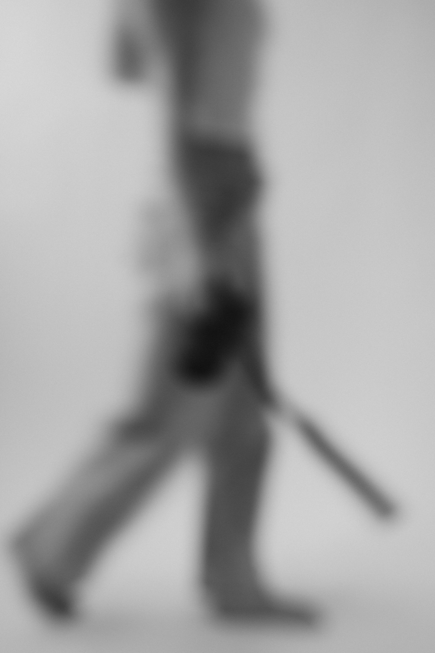 Black and white blurred image of person walking while carrying a camera with strap against gray background.