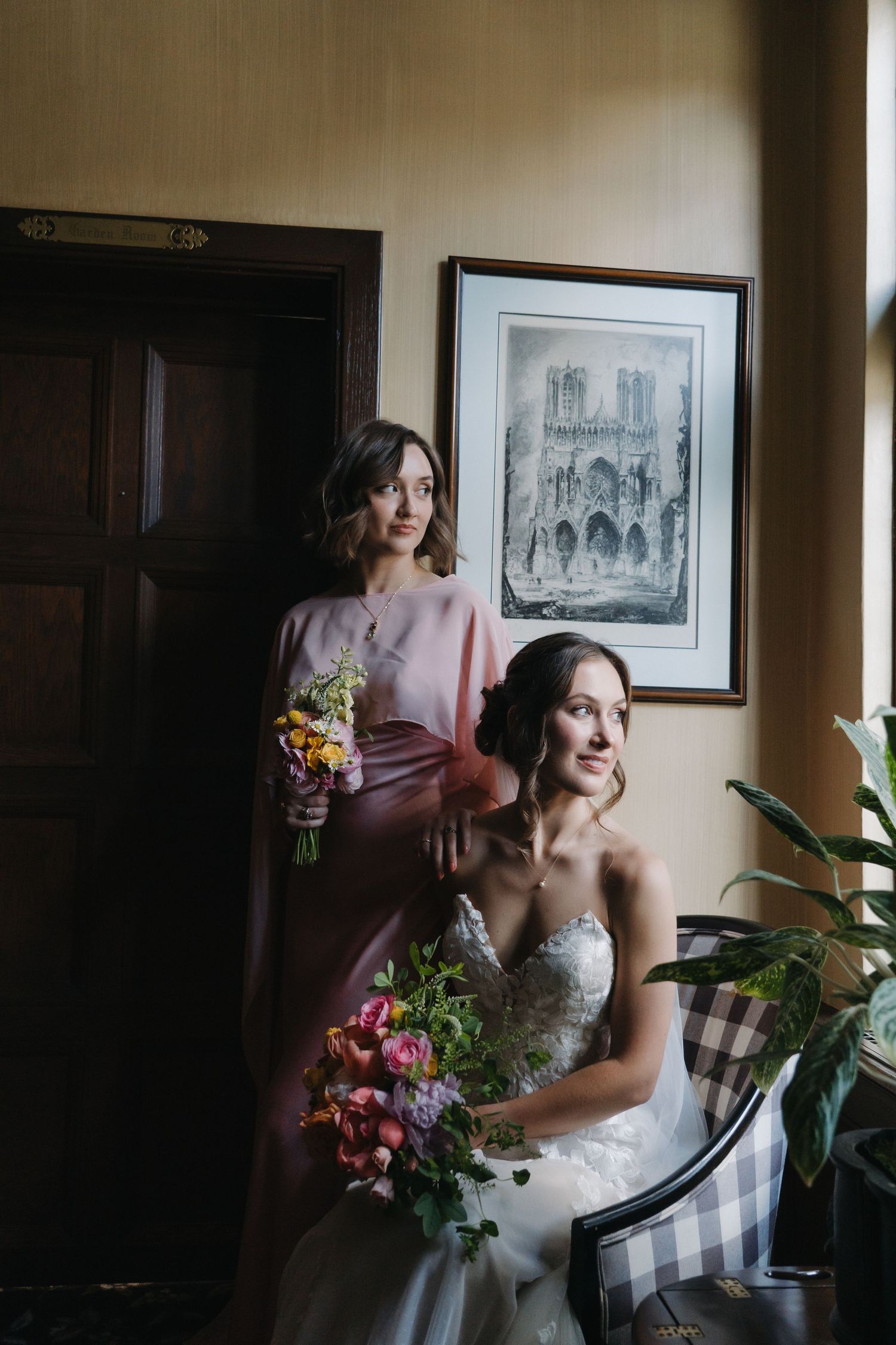 Bride in lace gown seated holding colorful bouquet with bridesmaid in pink dress standing beside her.