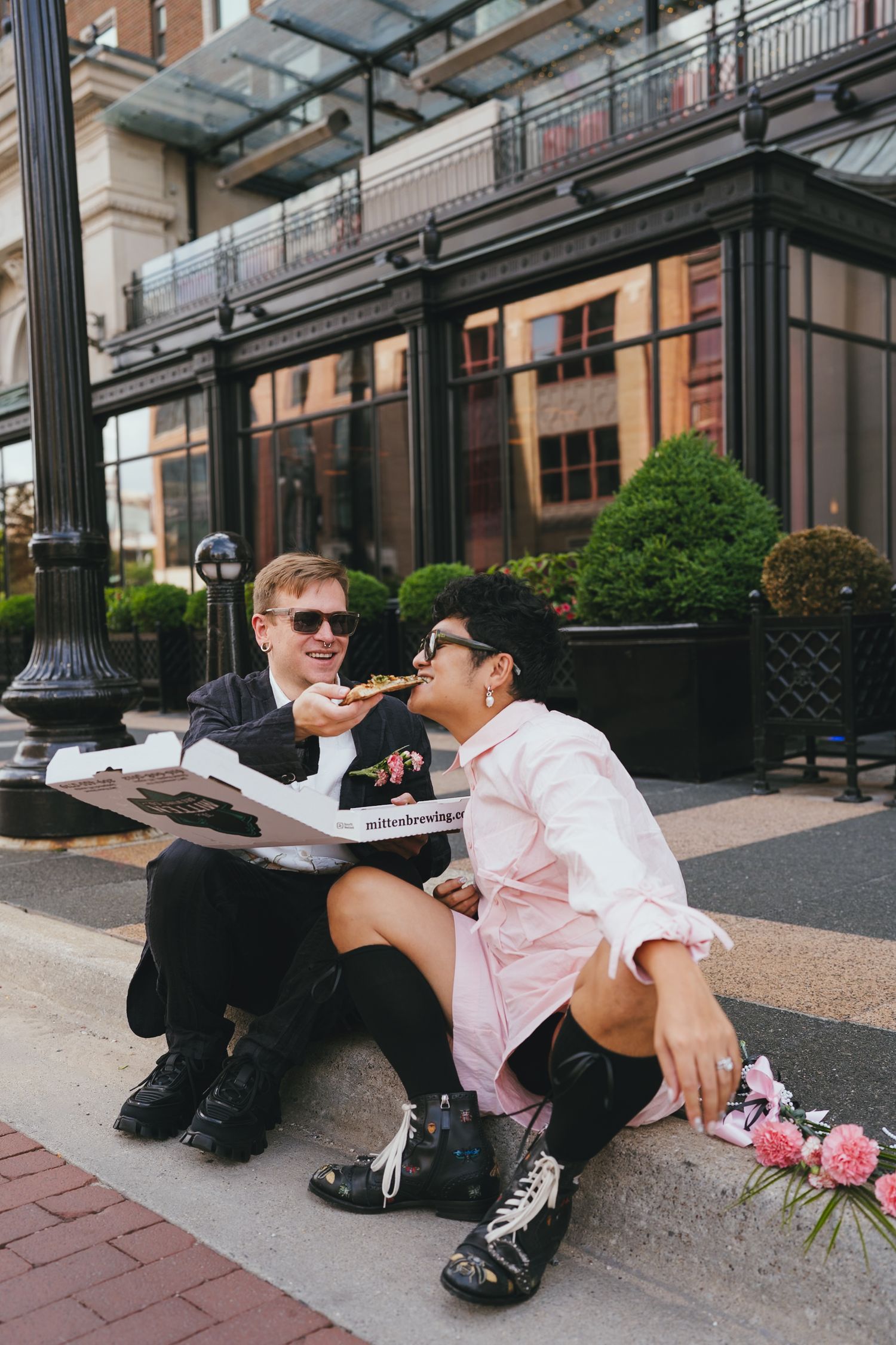 Couple in wedding attire sharing pizza on city steps, one feeding the other a slice.