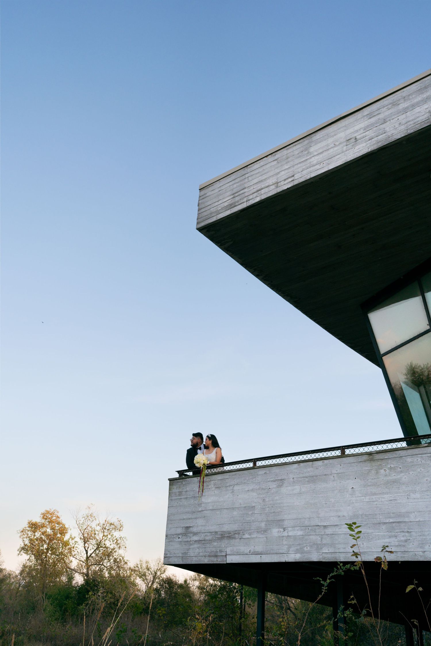 Wedding couple sharing romantic moment on modern concrete building balcony at sunset.