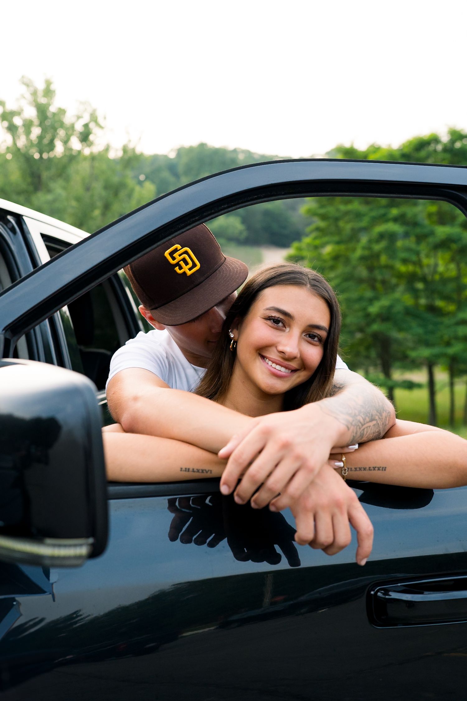Young woman smiling from car window while partner kisses her cheek in a scenic outdoor setting.