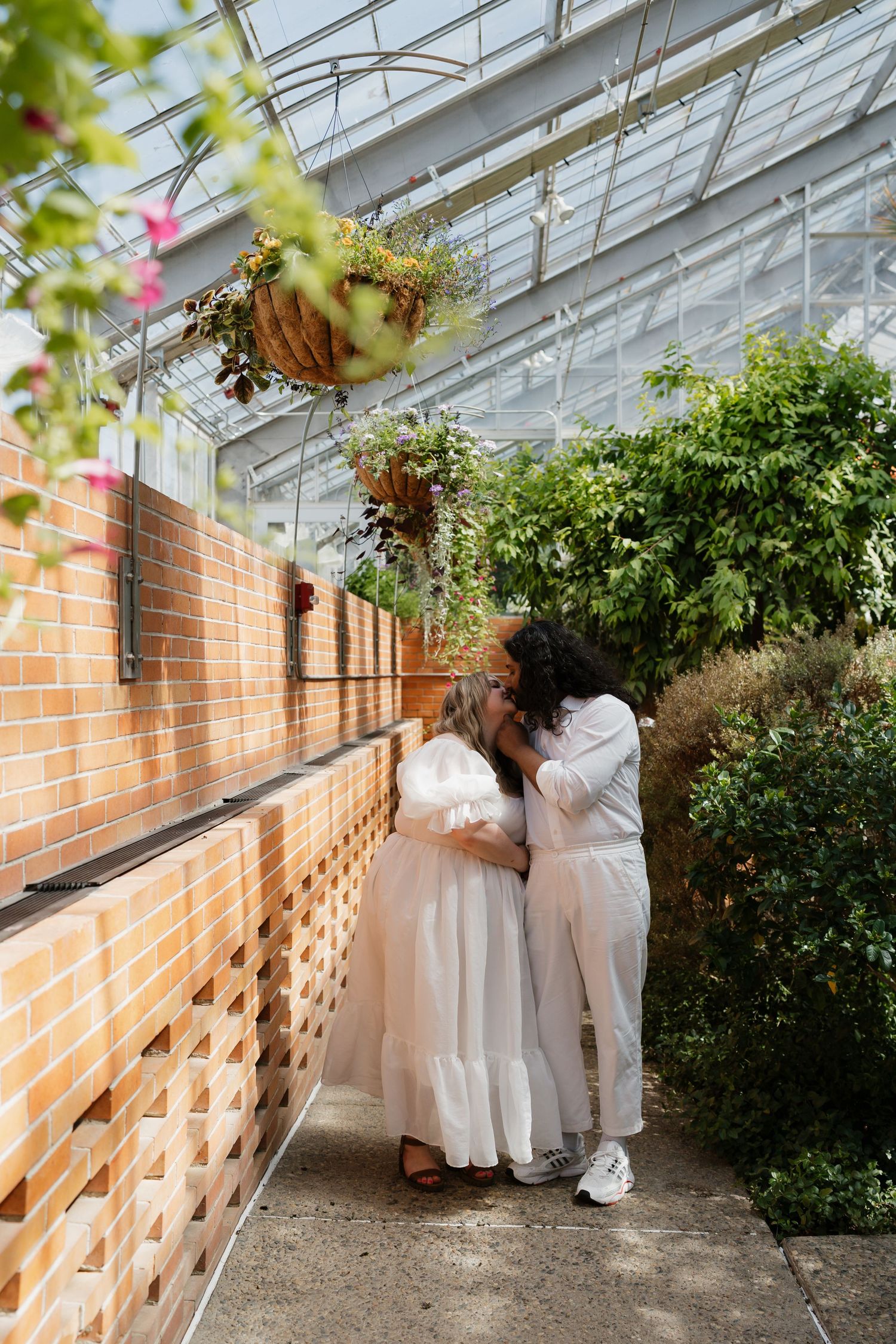 Couple sharing an intimate embrace in a greenhouse walkway with brick walls and hanging flowers.