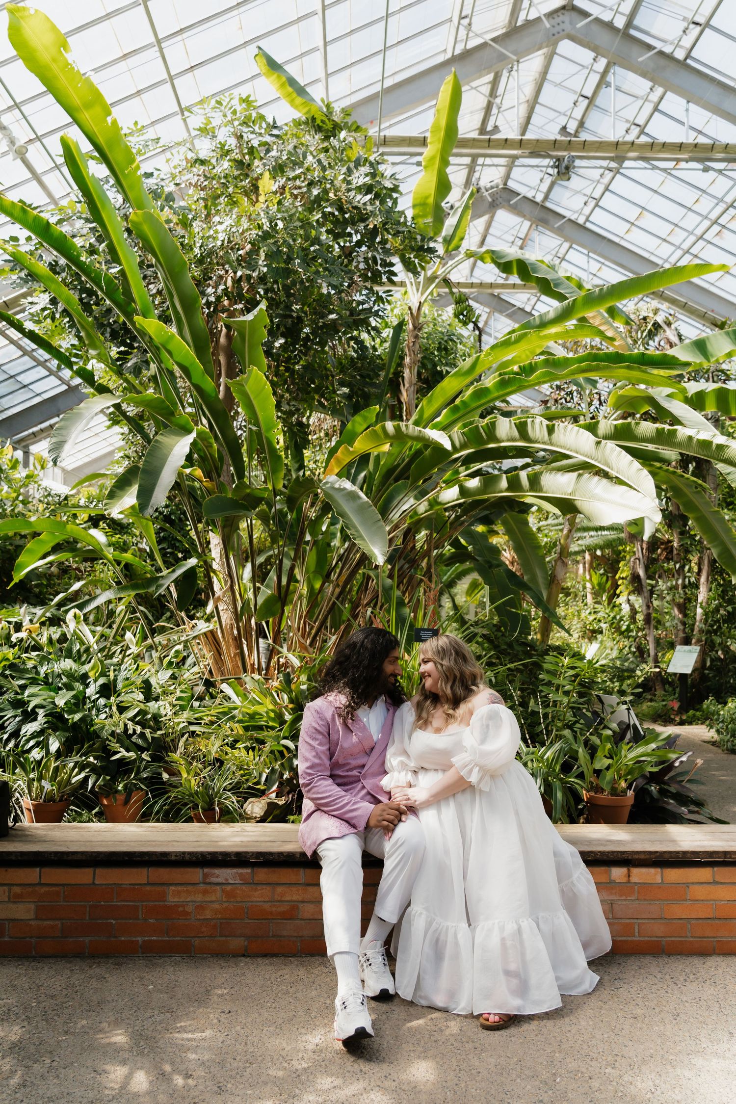Interracial couple in wedding attire sitting together in lush tropical greenhouse.