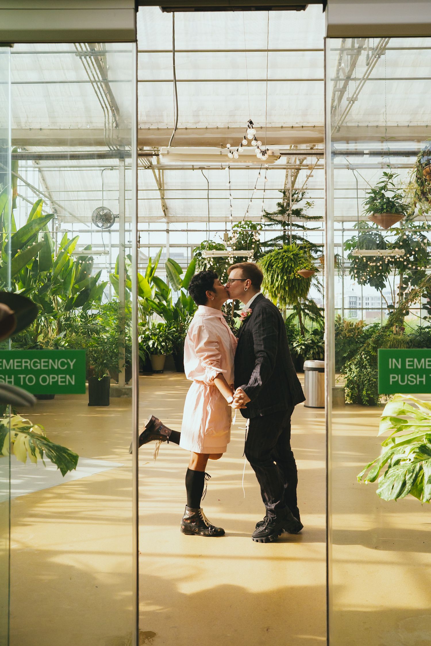 A couple shares a kiss framed by glass doors in a bright tropical greenhouse venue.