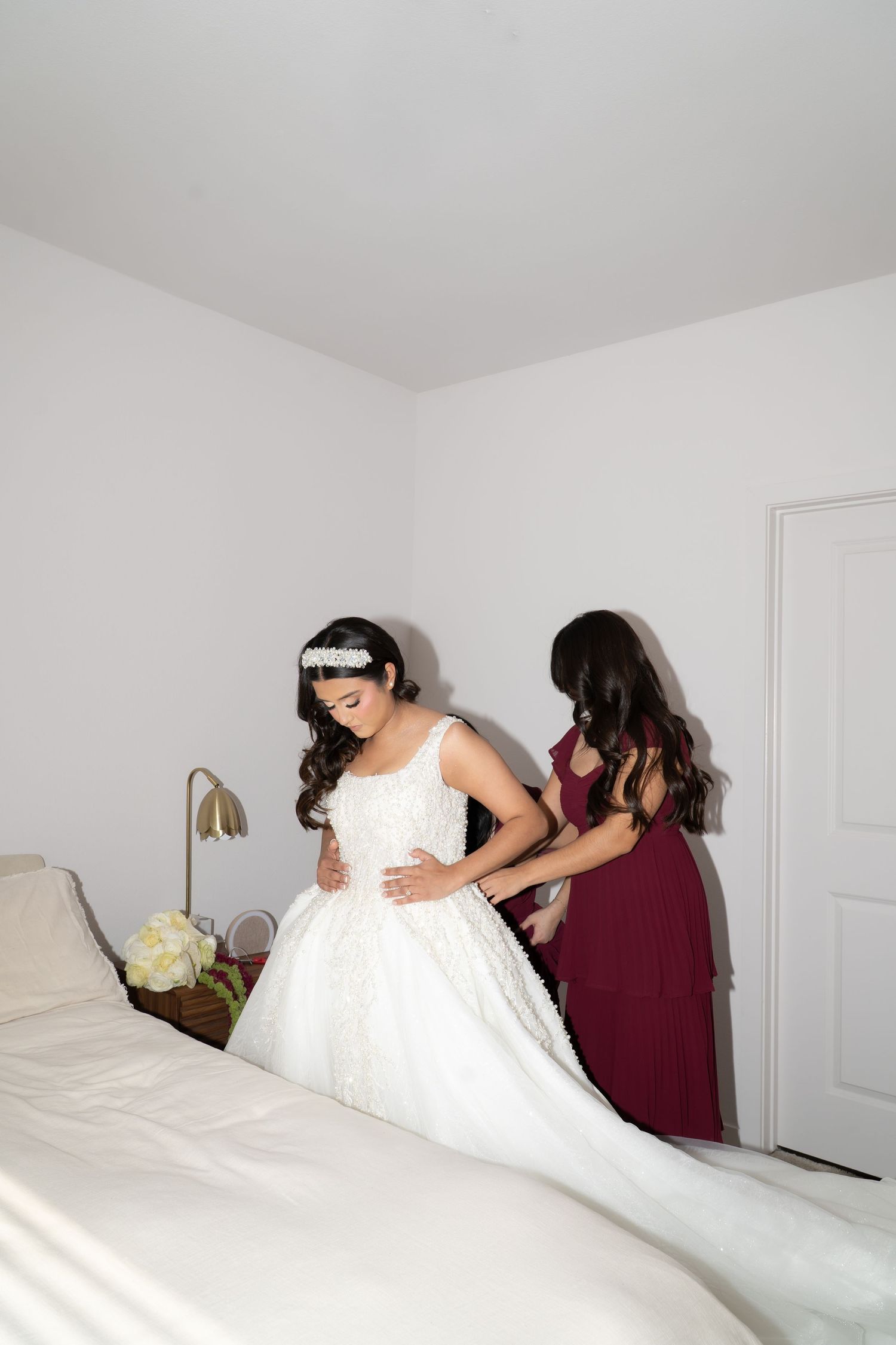 Bridesmaid in burgundy dress helping bride into elegant white lace ball gown wedding dress.