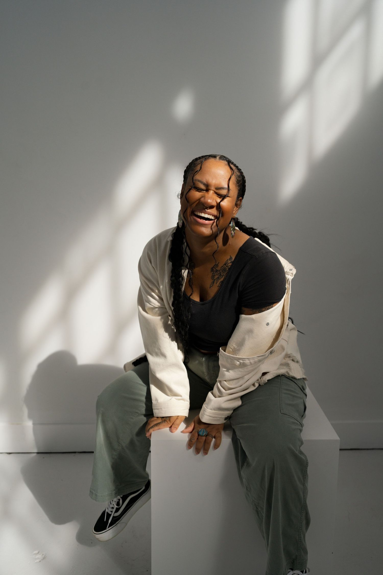 A joyful Black woman with braids laughs while sitting on a white cube in a bright sunlit studio.