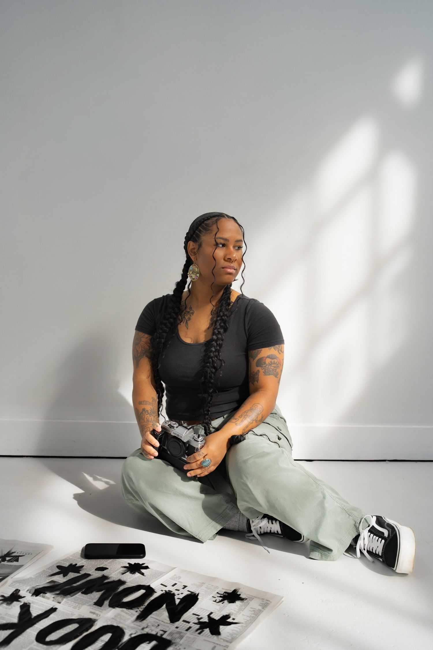 A Black woman with long braids sits on a floor in a black top, sage green pants, and Vans sneakers, holding a camera.