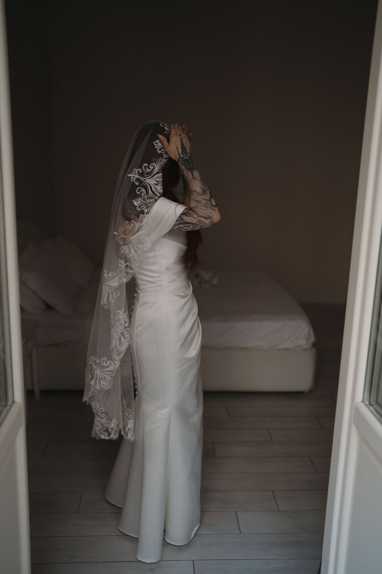 Bride in elegant white satin gown and lace veil standing in doorway of softly lit bedroom.