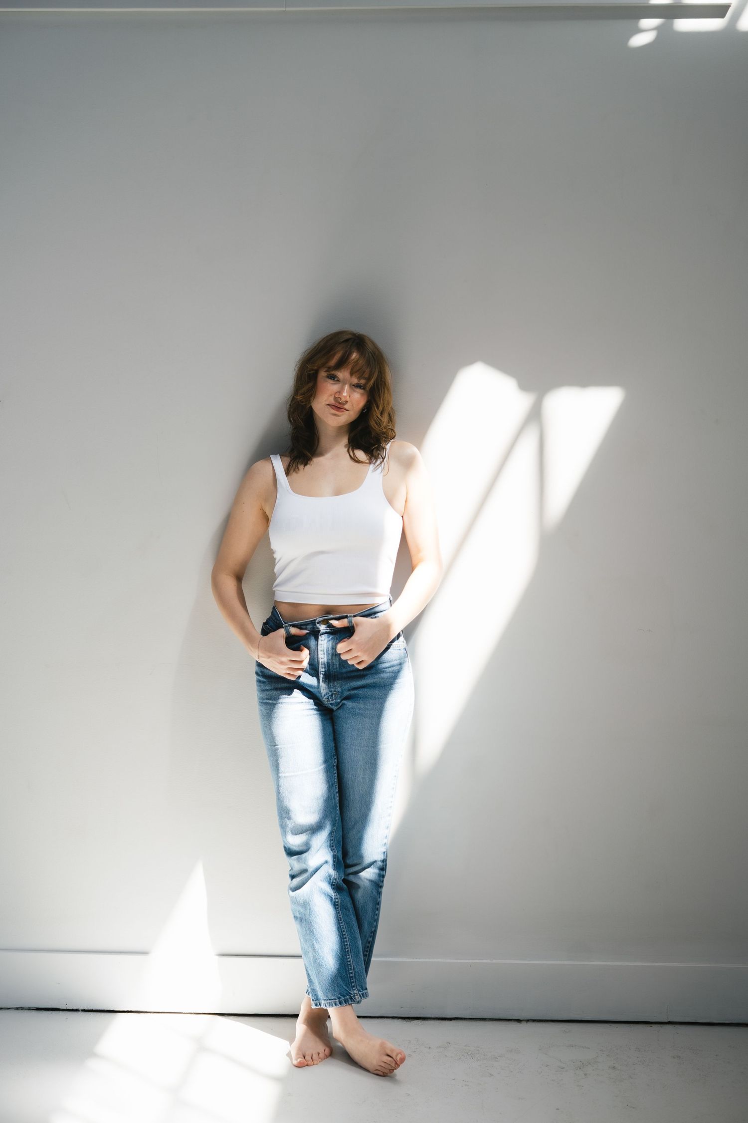 Woman in white crop tank top and blue straight-leg jeans standing barefoot in sunlit white studio.