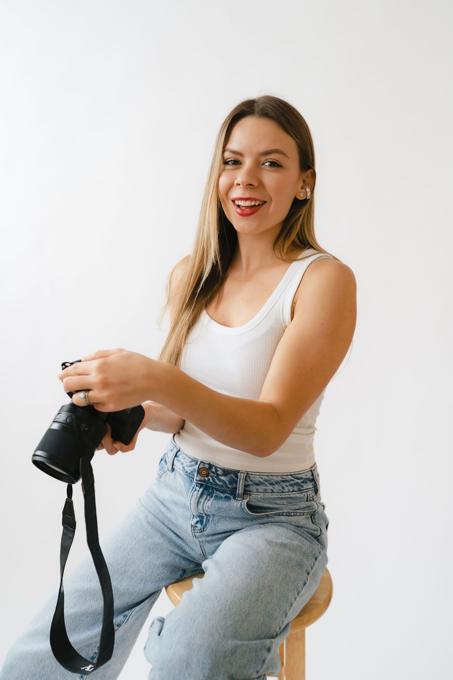 Happy young woman in white tank top and jeans holding a Sony camera while seated on a wooden stool.