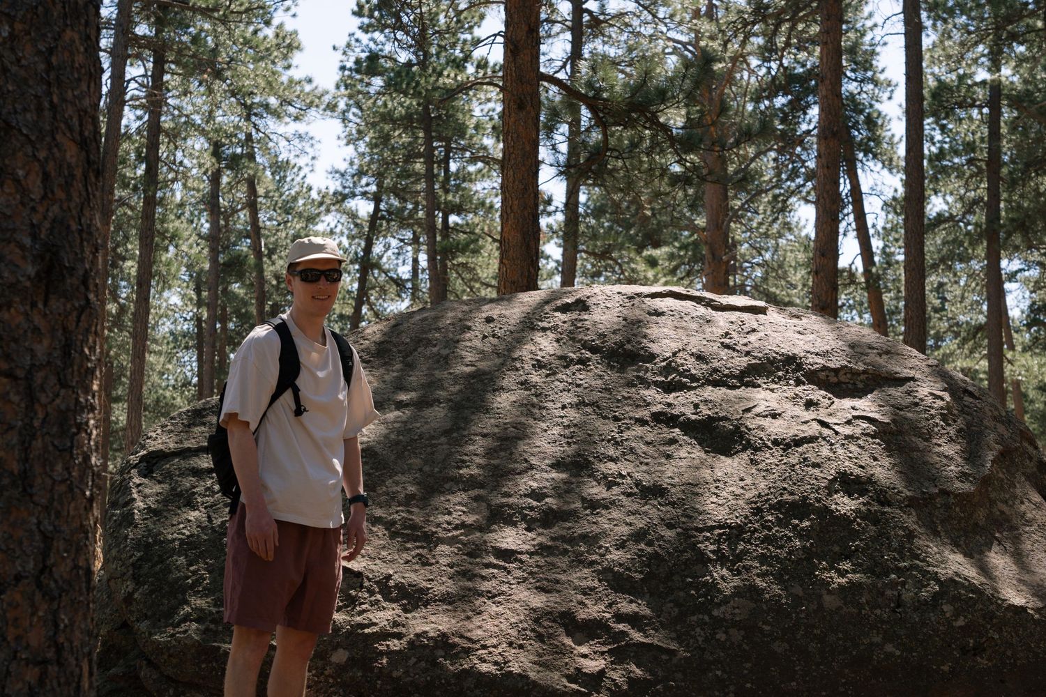 A hiker in a white shirt and hat stands beside a large granite boulder in a pine forest.
