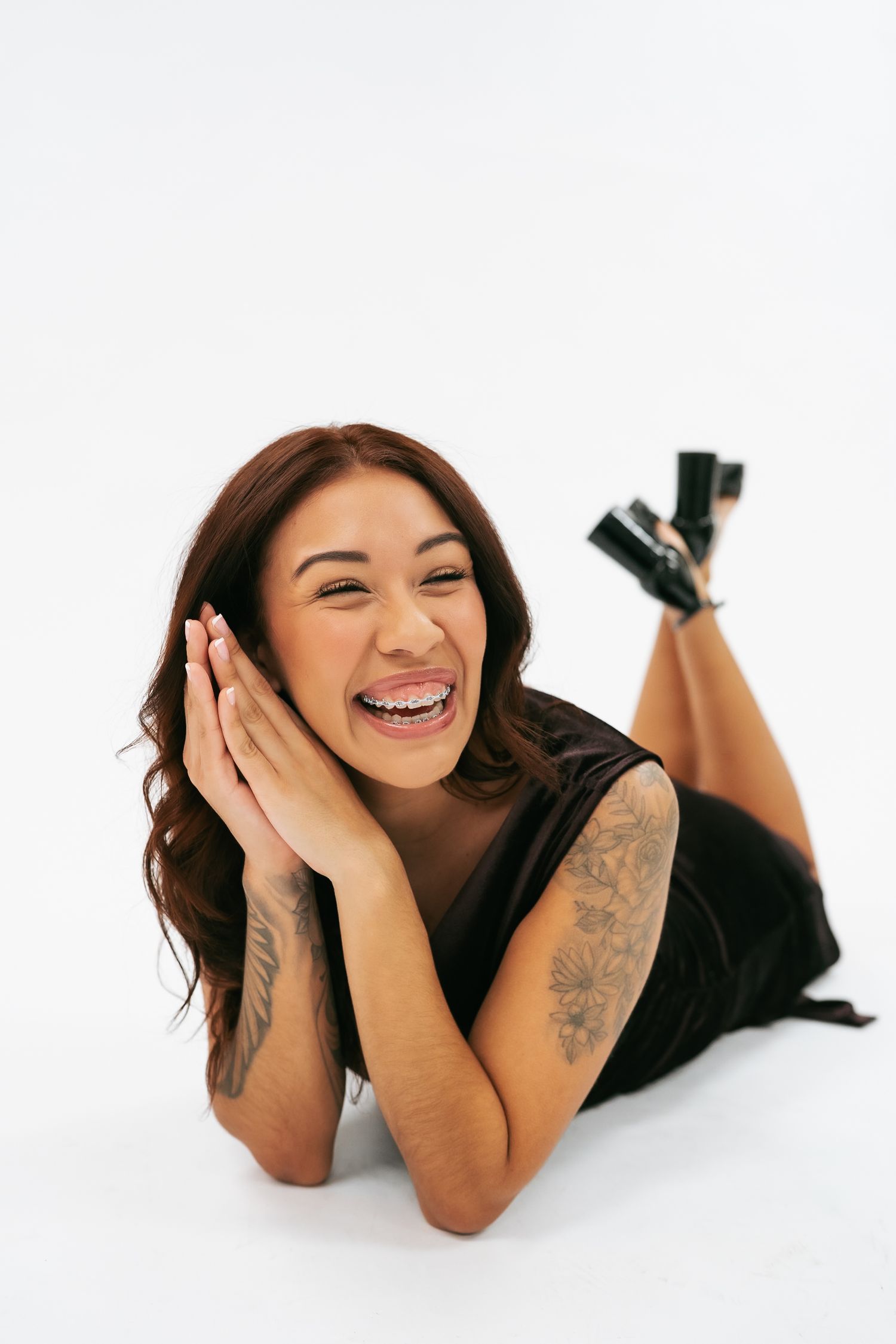 Smiling tattooed woman in black dress lying on white studio floor laughing with hands near face.