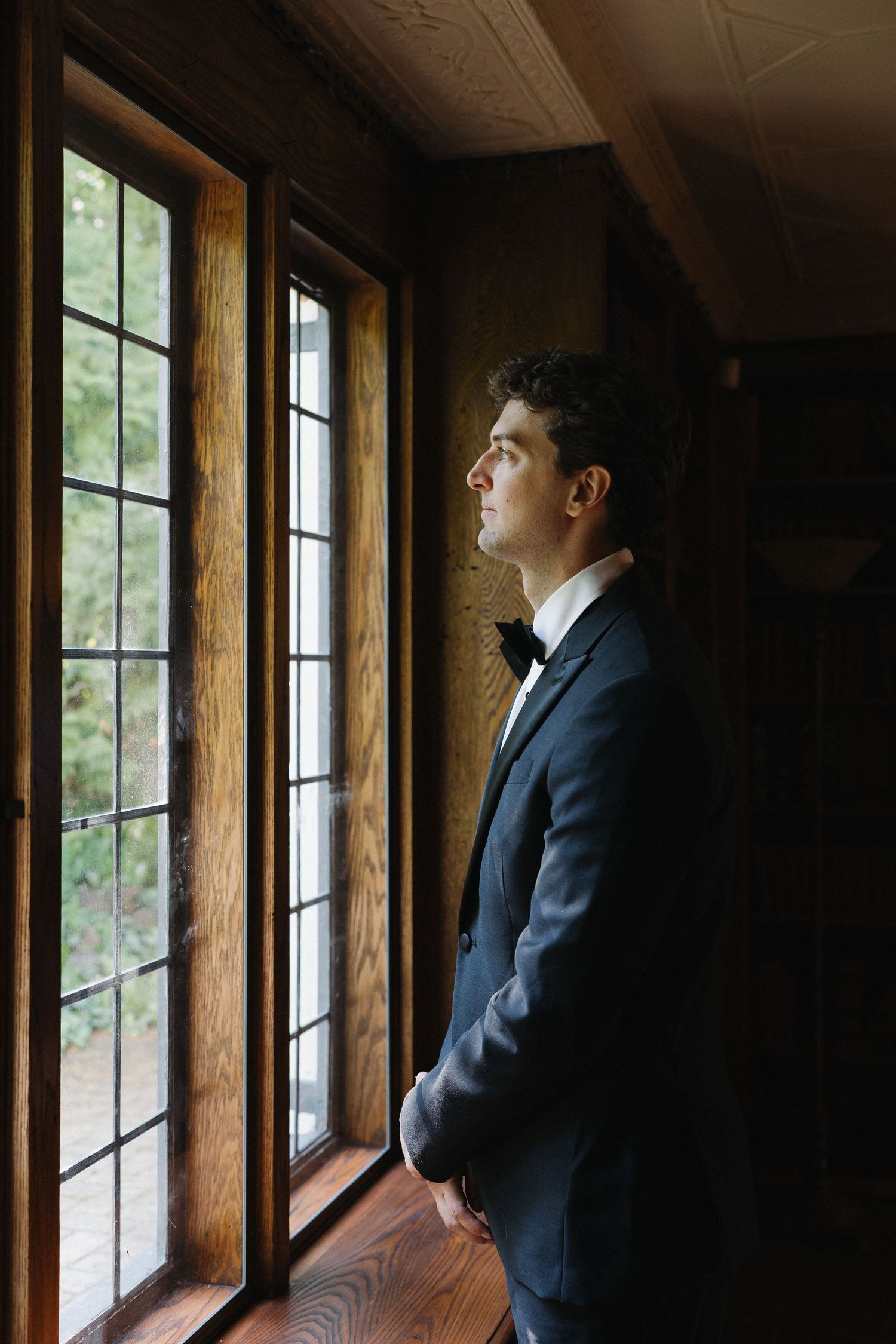 Handsome groom in black tuxedo gazing thoughtfully out leaded glass windows before wedding ceremony.