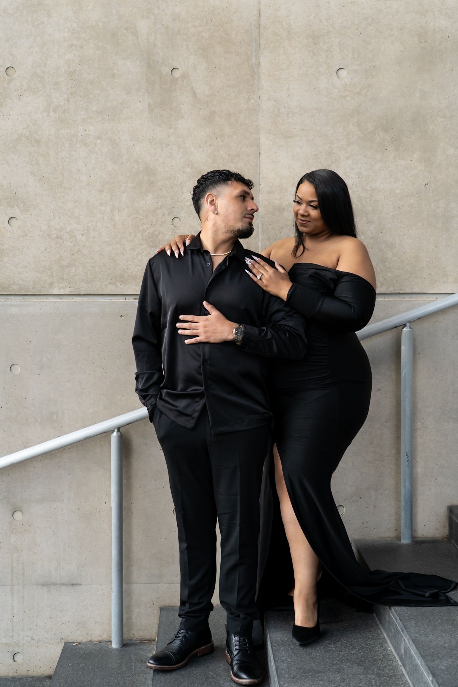 Elegant couple in matching black formal attire posing together against a concrete wall.