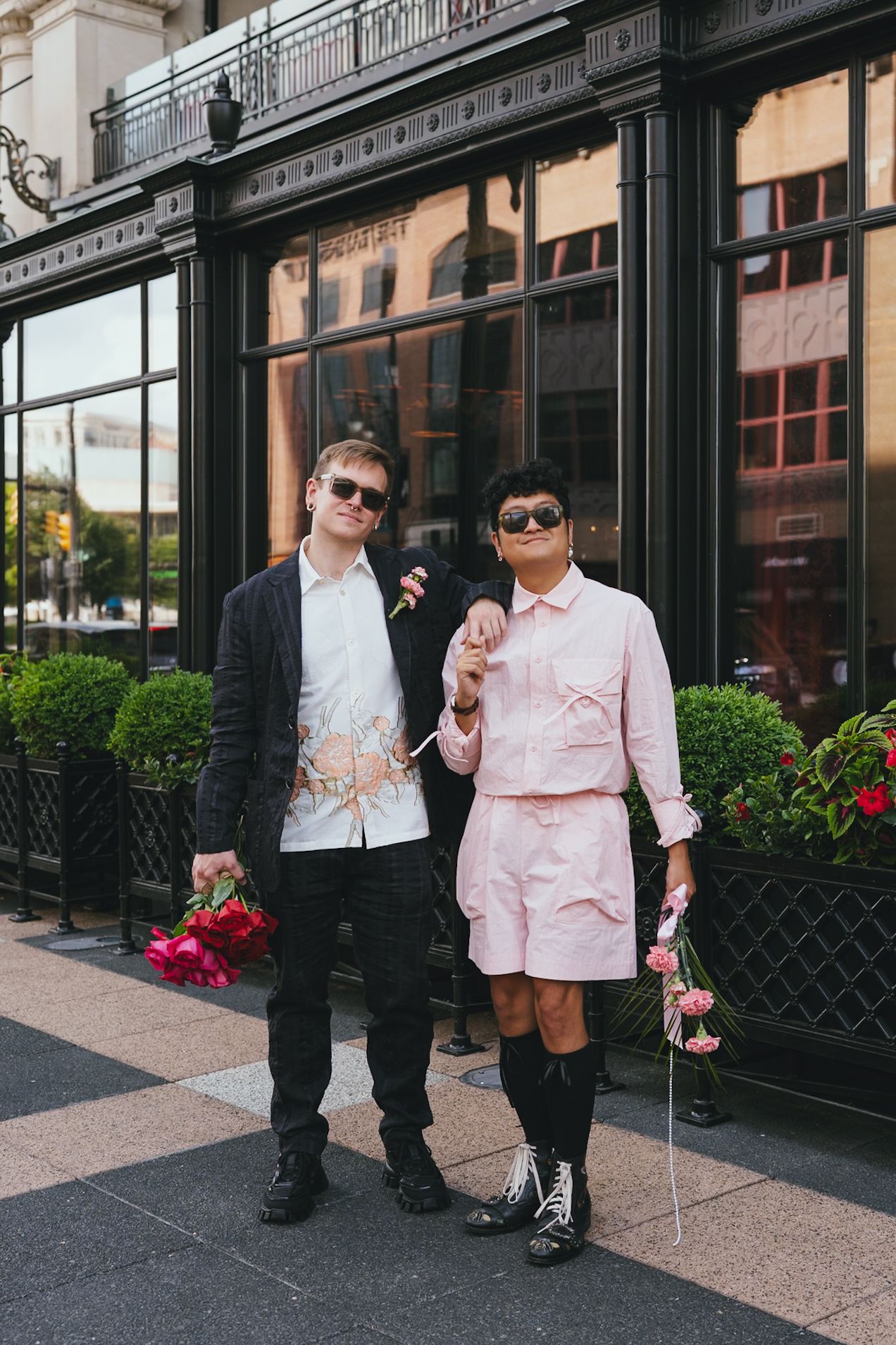 Stylish newlywed couple in sunglasses holding pink flowers outside elegant urban building.